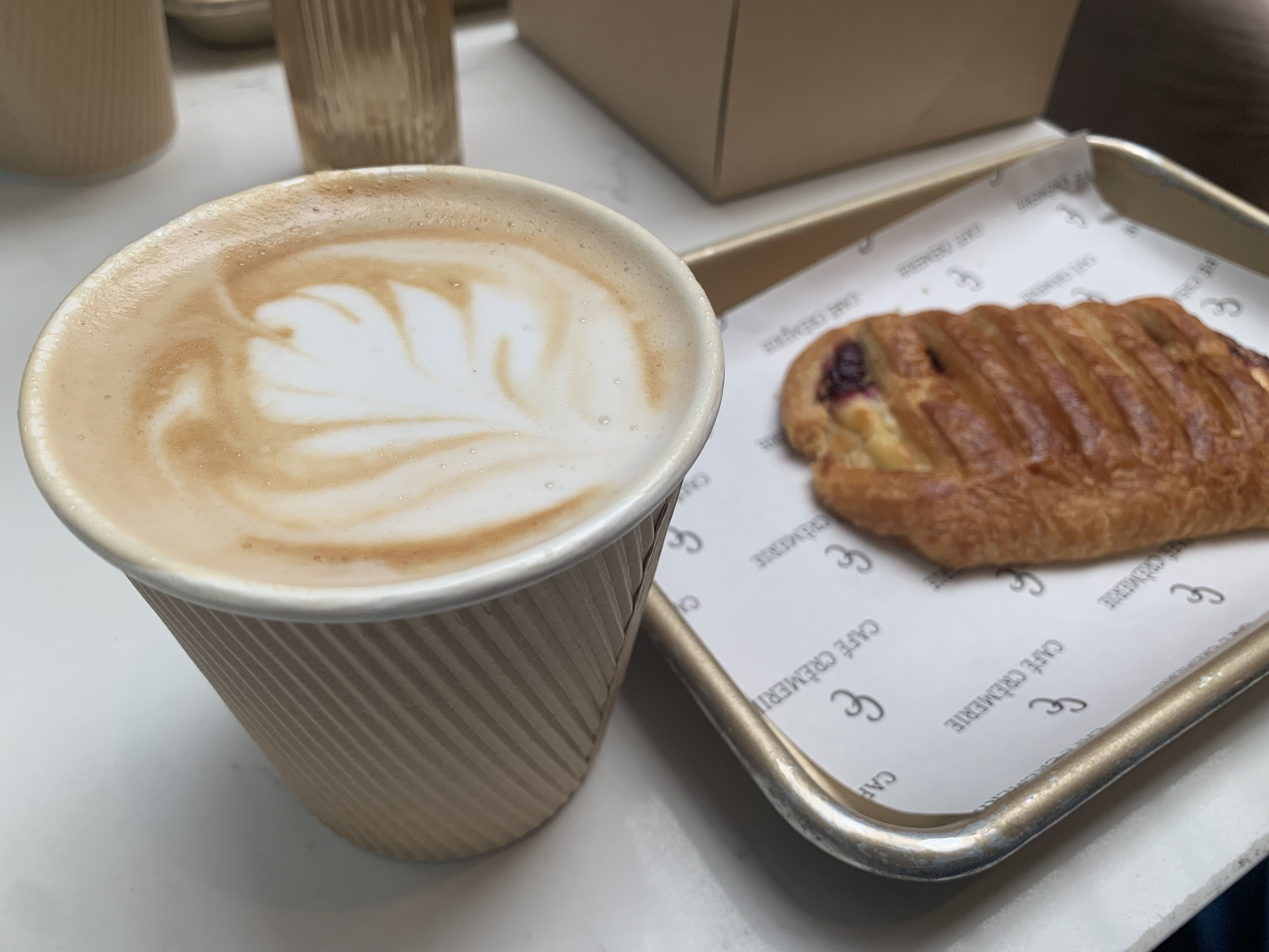 Close-up of a latte with leaf latte art in a brown paper cup, beside a blueberry Danish on a metal tray lined with printed paper.