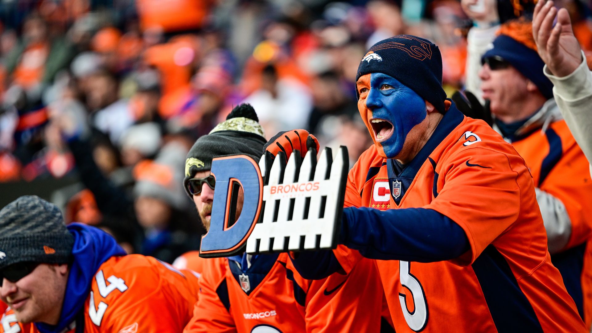 A Broncos fan with blue and orange face paint hold a "defense" sign and cheers 