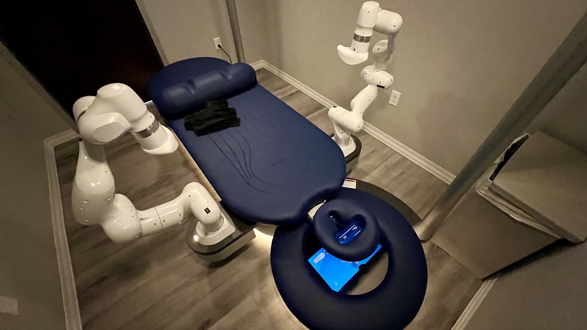 Blue cushioned massage table with two white robotic arms on the sides, a face rest with a tablet underneath it, and folded black towels on the table in a neutral-toned room.