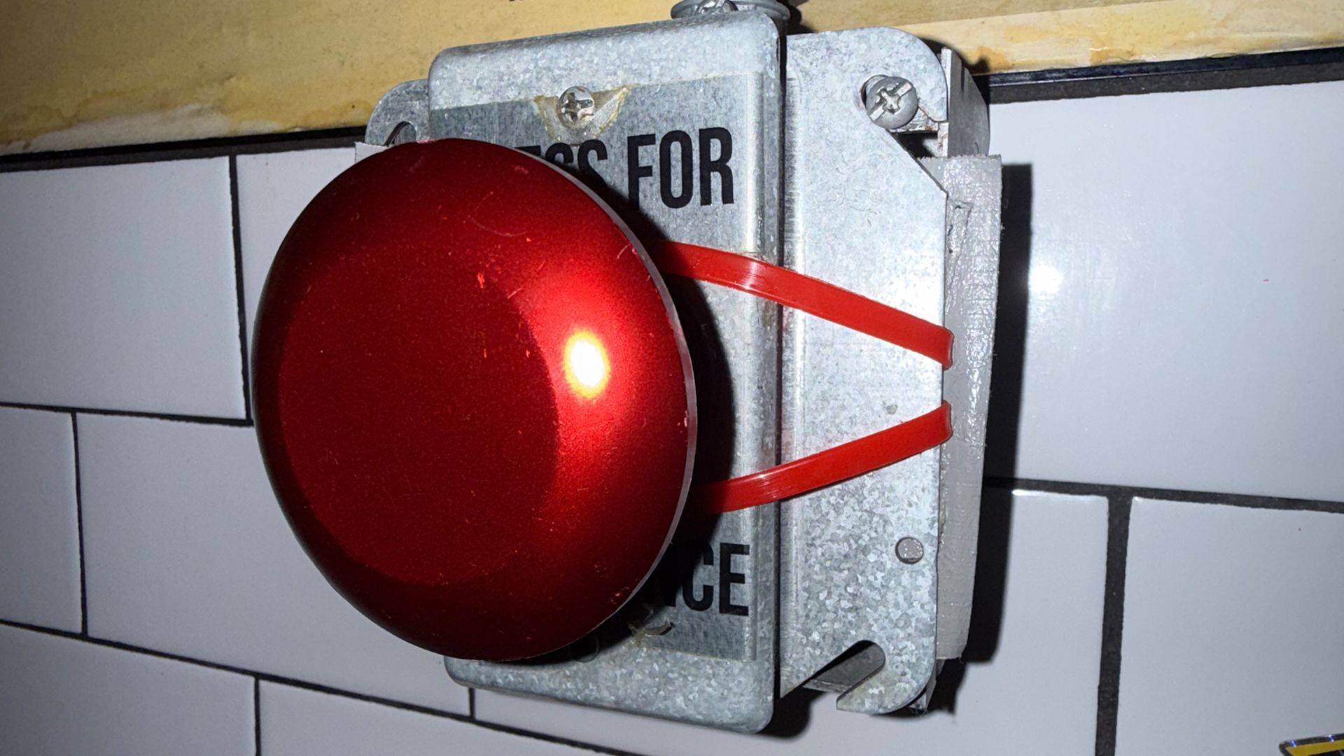 A large red emergency stop button mounted on a gray metal switch box secured to a tiled wall; two red straps wrap around the box, with partial letters 'FOR' visible on the label.