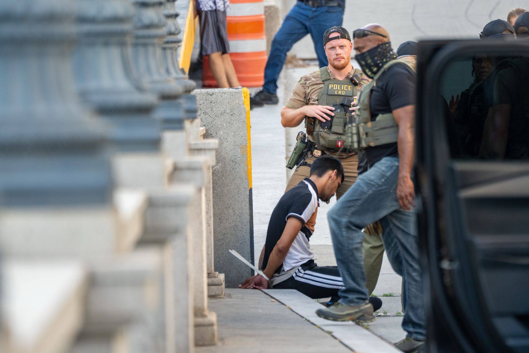 A man in a black and white shirt sits handcuffed on the sidewalk near concrete barriers while police officers, one wearing a vest labeled "POLICE ERO," stand nearby.