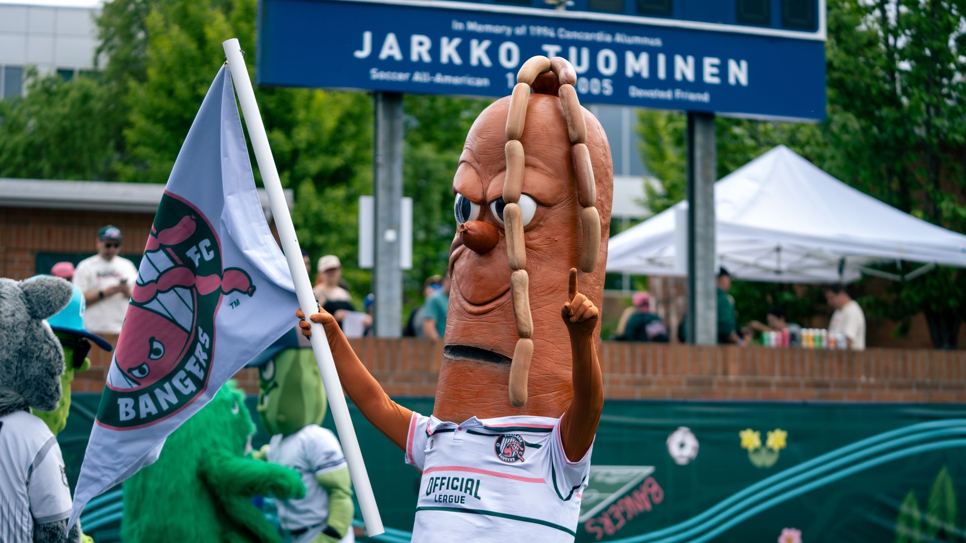 Mascot dressed as a hot dog with sausages on top, holding a white flag with a sausage logo, at a sports event with other mascots and people in the background.