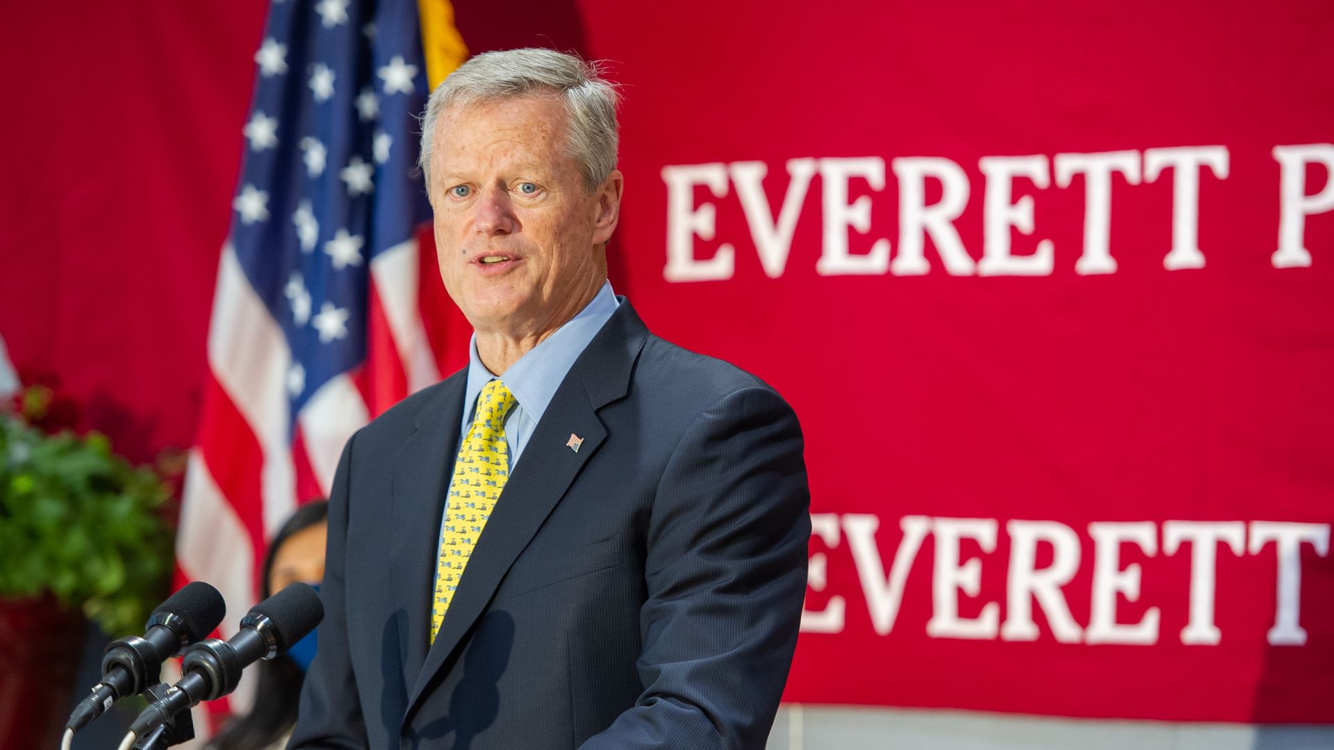 Photo of Charlie Baker in a suit speaking from a podium with a red wall and the American flag behind him 