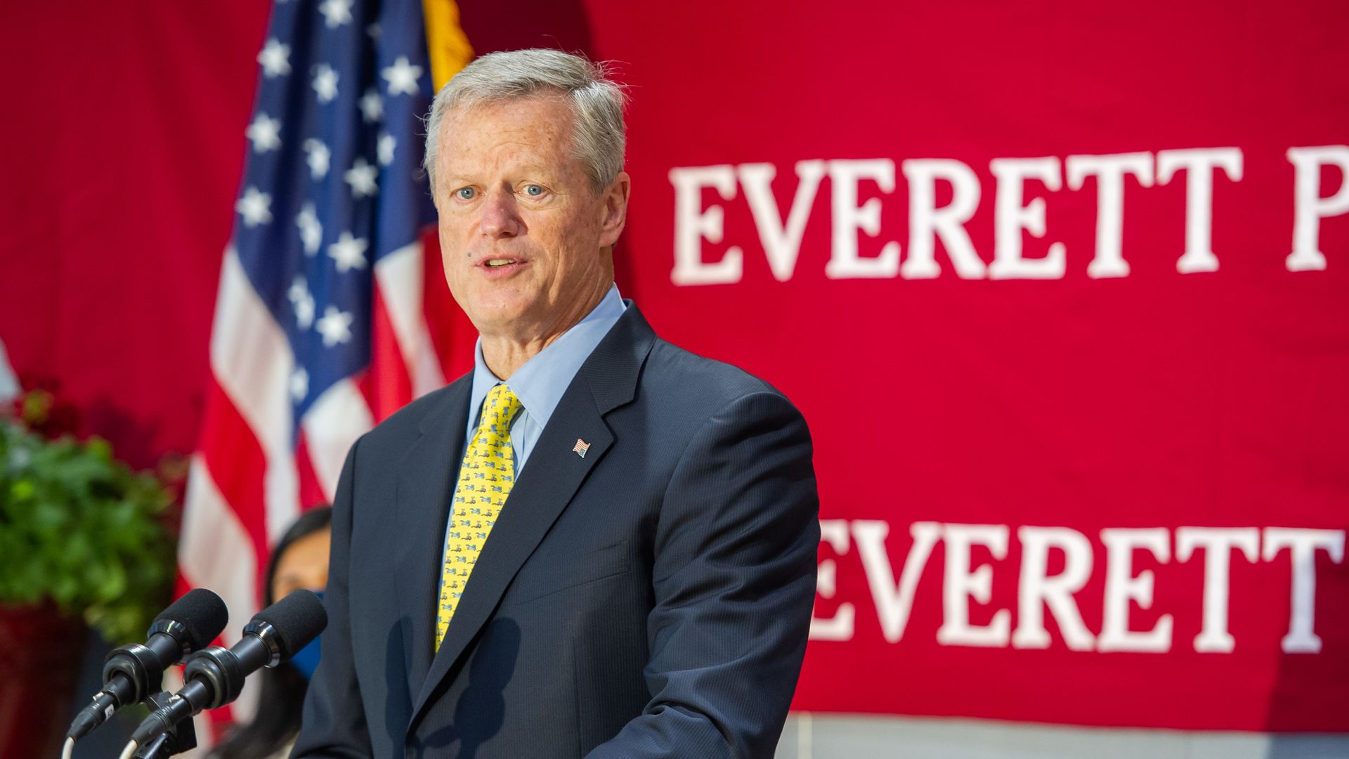 Photo of Charlie Baker in a suit speaking from a podium with a red wall and the American flag behind him
