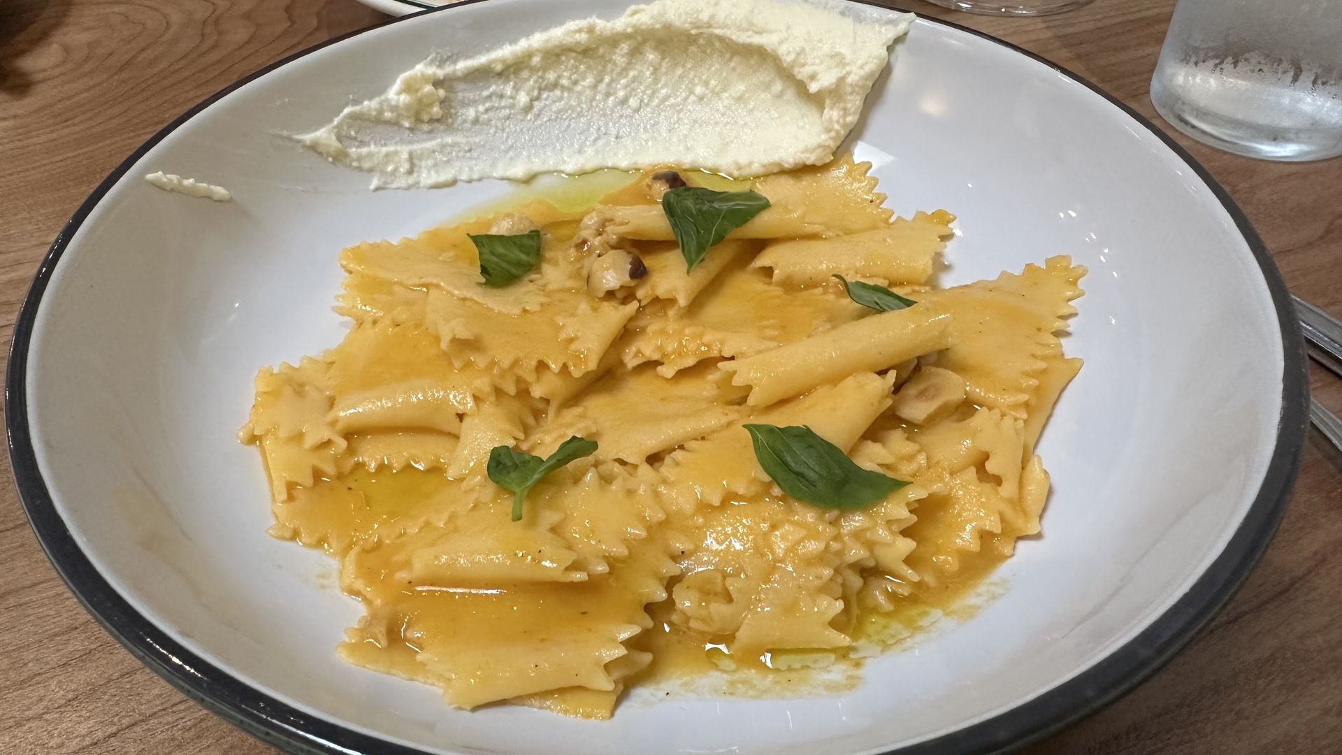 Pasta in cream sauce on a white plate, garnished with fresh basil; a dollop of white ricotta rests at the top edge. Wooden table and a glass nearby.