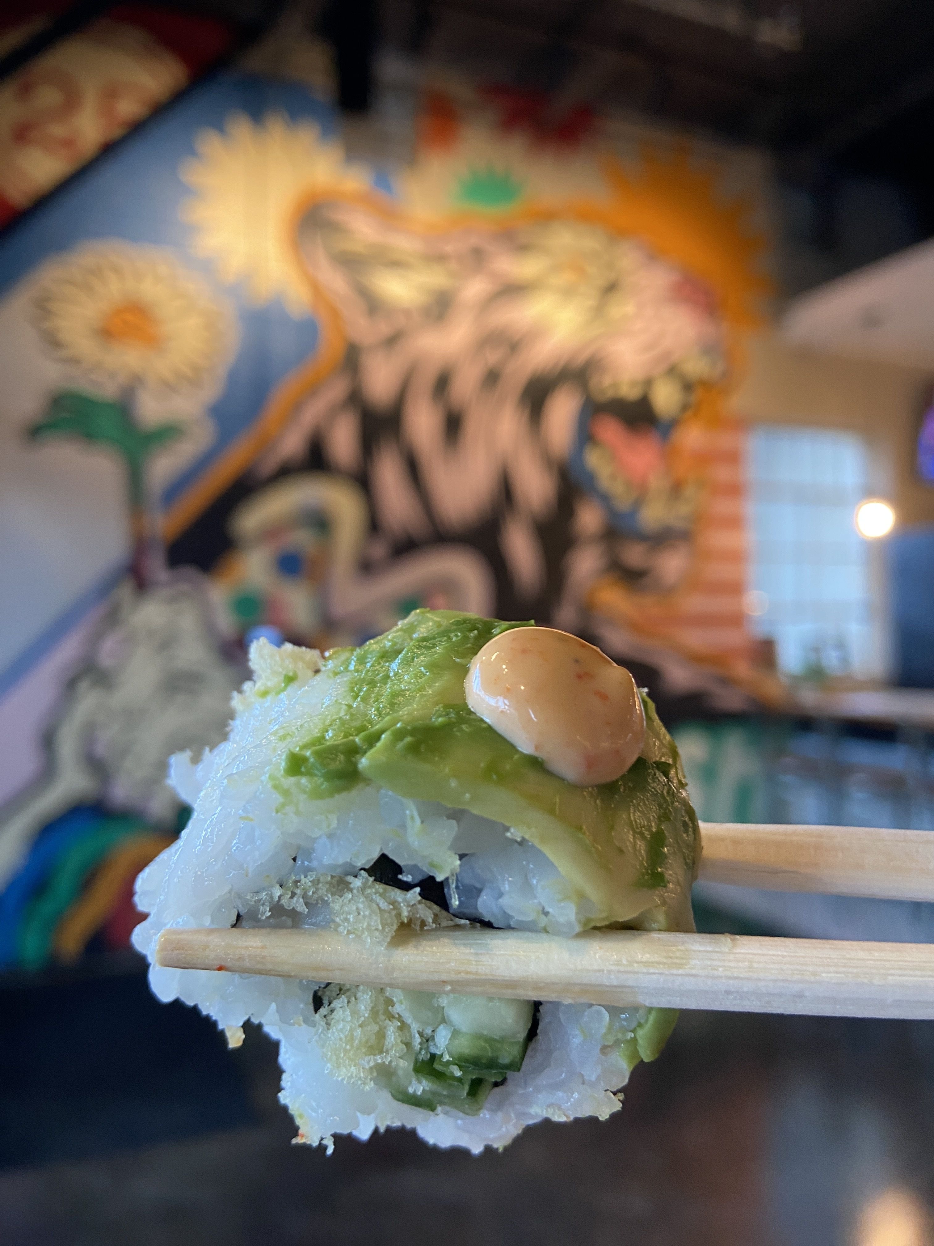 Close-up of a sushi roll held by chopsticks with avocado slice and spicy mayo on top, colorful wall mural featuring a roaring tiger and sunflowers in the background.
