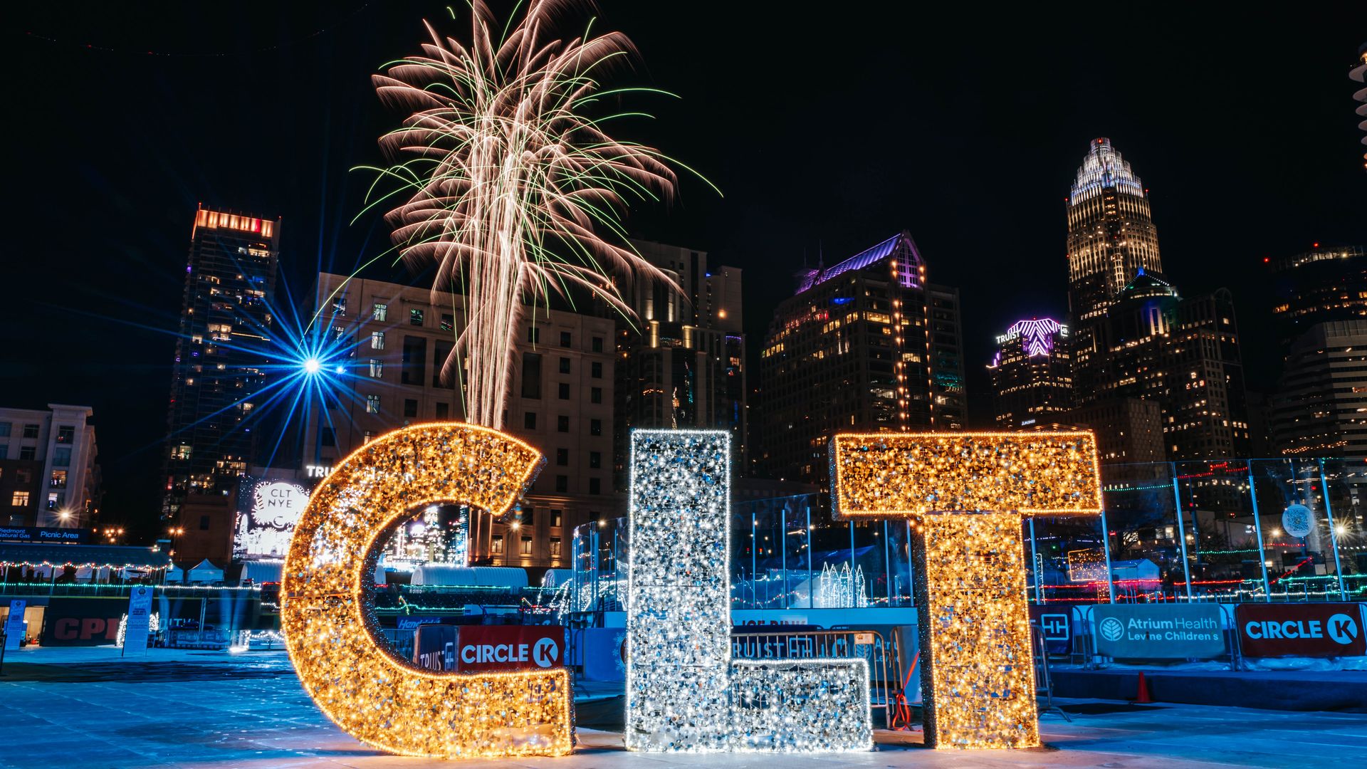 Large illuminated letters "CLT" in orange and white lights with fireworks above, set against a nighttime city skyline with lit buildings and a clear sky.