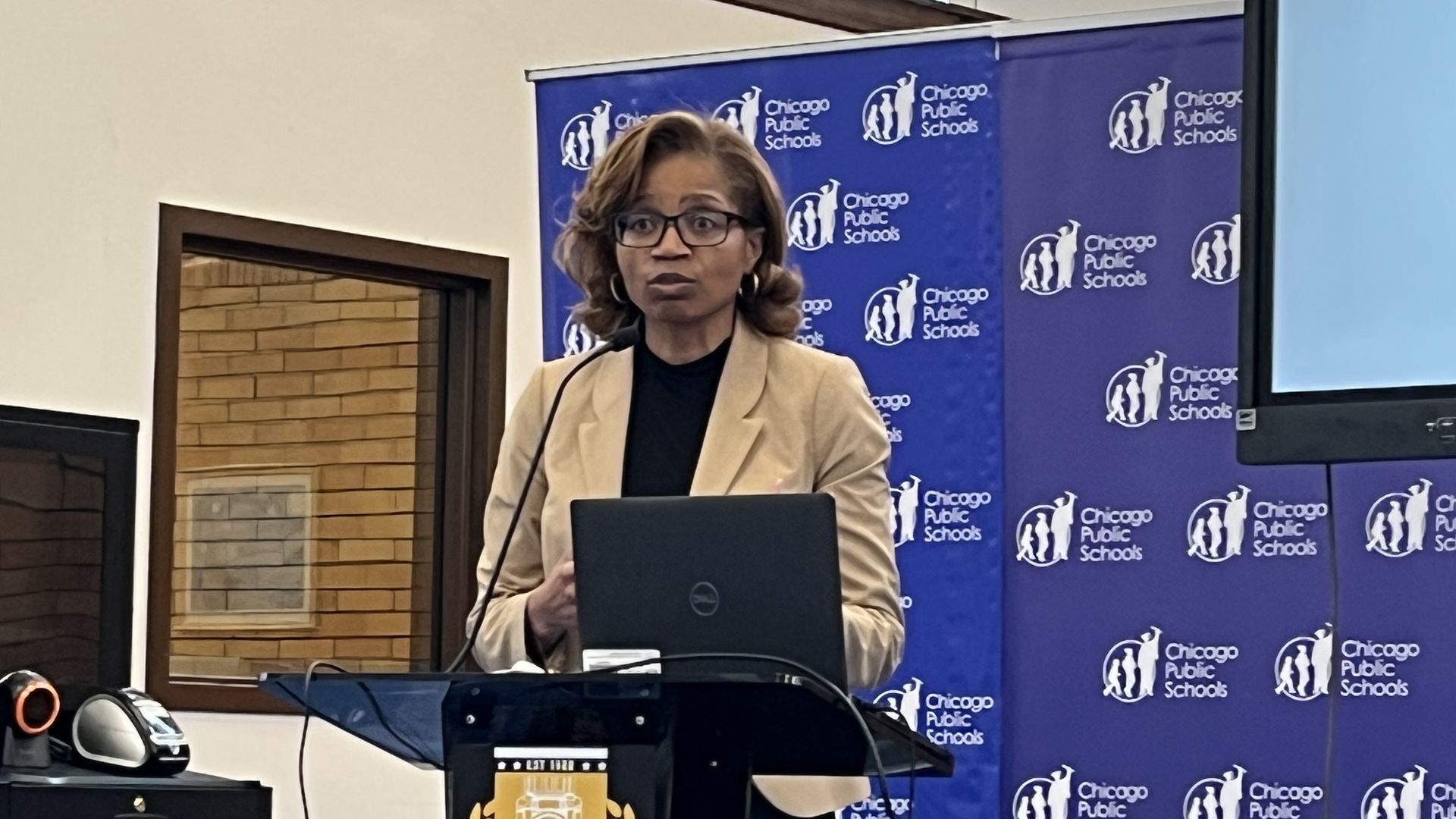 A woman in a tan blazer, black top, and glasses speaks at a podium with a laptop. A blue backdrop repeats Chicago Public Schools logos; the podium bears a Roosevelt High School crest.