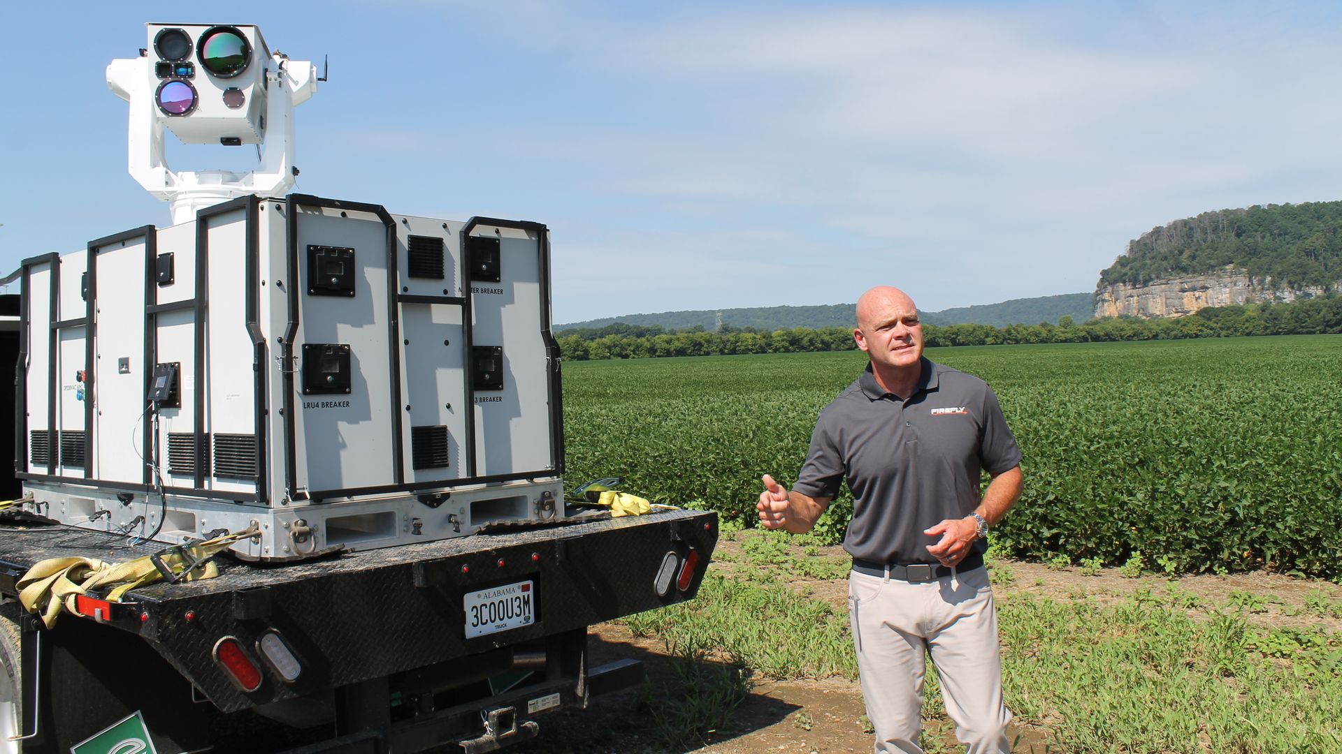 Large white mobile equipment unit on a trailer with multiple lenses; a man in a gray polo stands in a green field under a blue sky, with a distant hill/cliff in the background.
