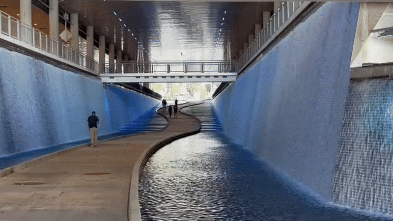 Indoor walkway with a winding path between two blue walls where water cascades down like waterfalls underneath the David L. Lawrence Convention Center in Pittsburgh, with people walking and a ceiling reflecting the water's movement.