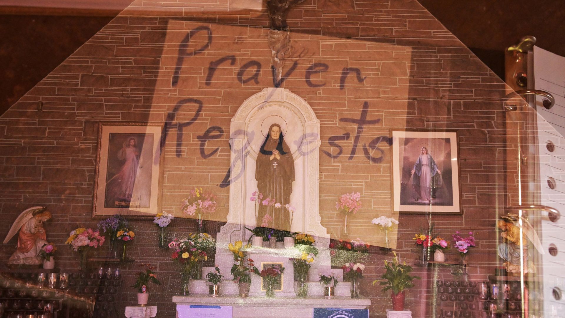 A shrine featuring a small statute of a woman in dark robes, with hands in prayer, and a Christian cross necklace. Around her har many bouquets of flowers, two other images depicting Christ and the Virgin Mary hung on a brick wall. 