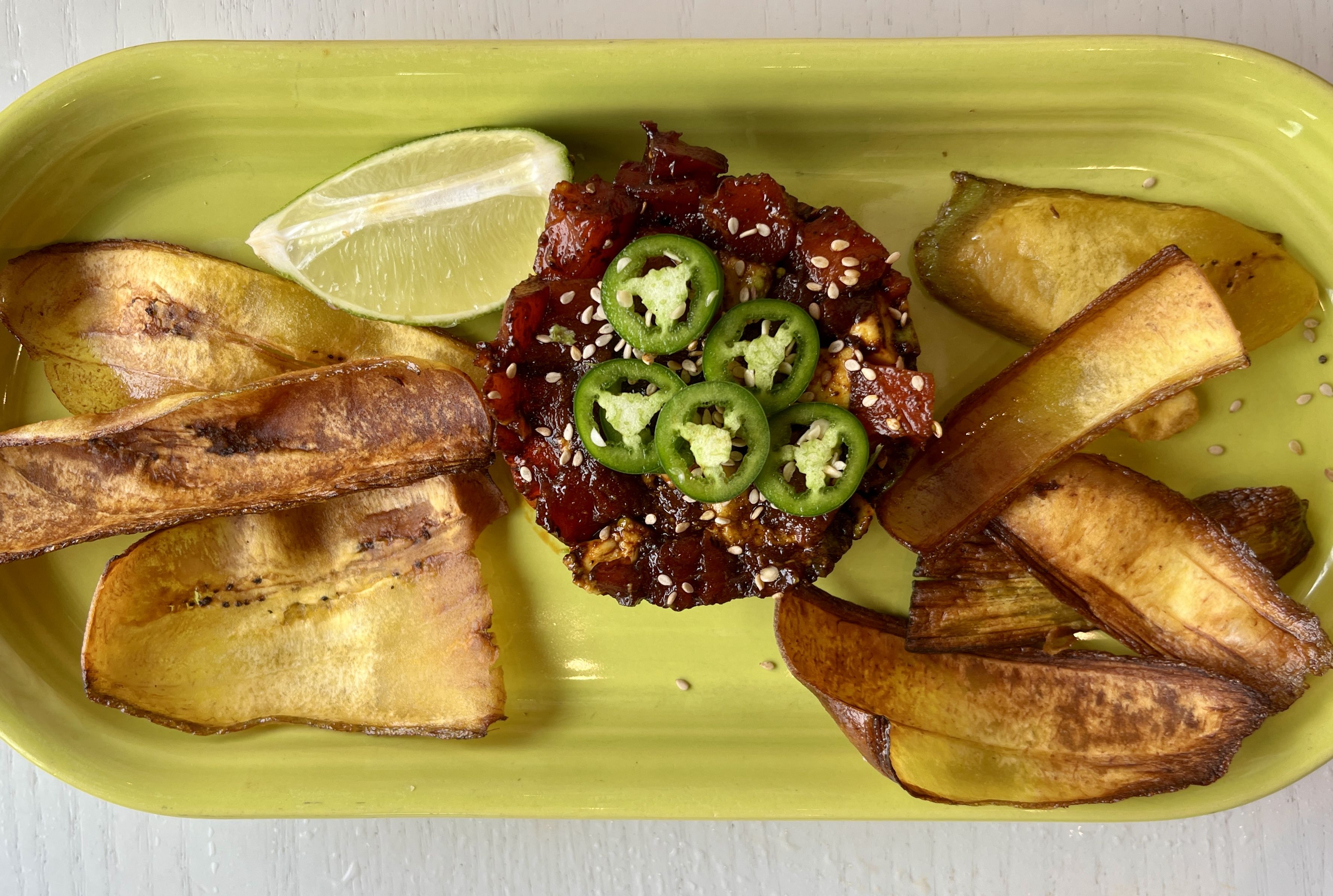Green rectangular plate with a serving of diced marinated meat topped with sliced green jalapeños and sesame seeds, accompanied by plantain chips and a lime wedge.