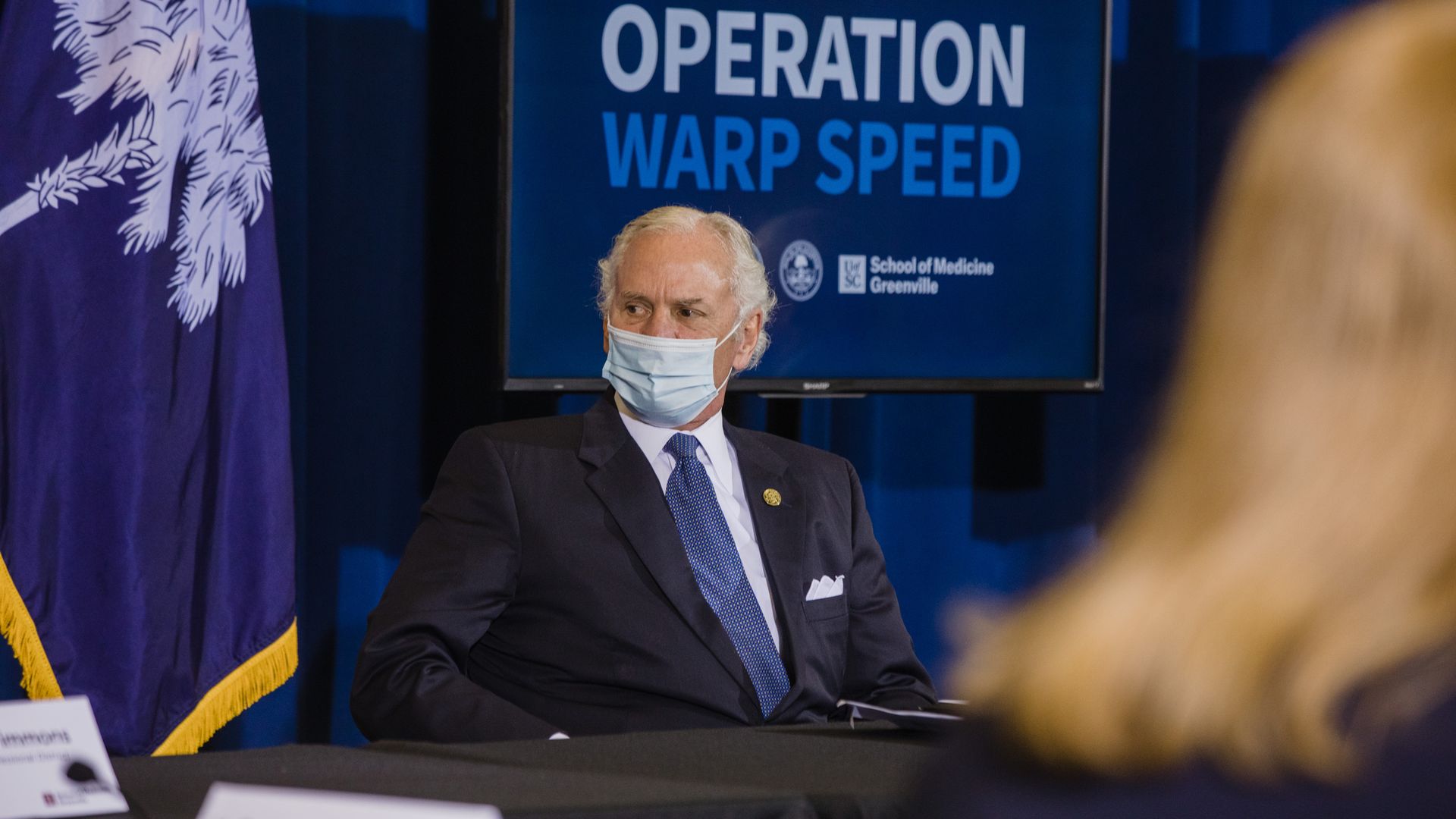 Photo of a masked Henry McMaster sitting in front of a sign that reads "Operation Warp Speed"