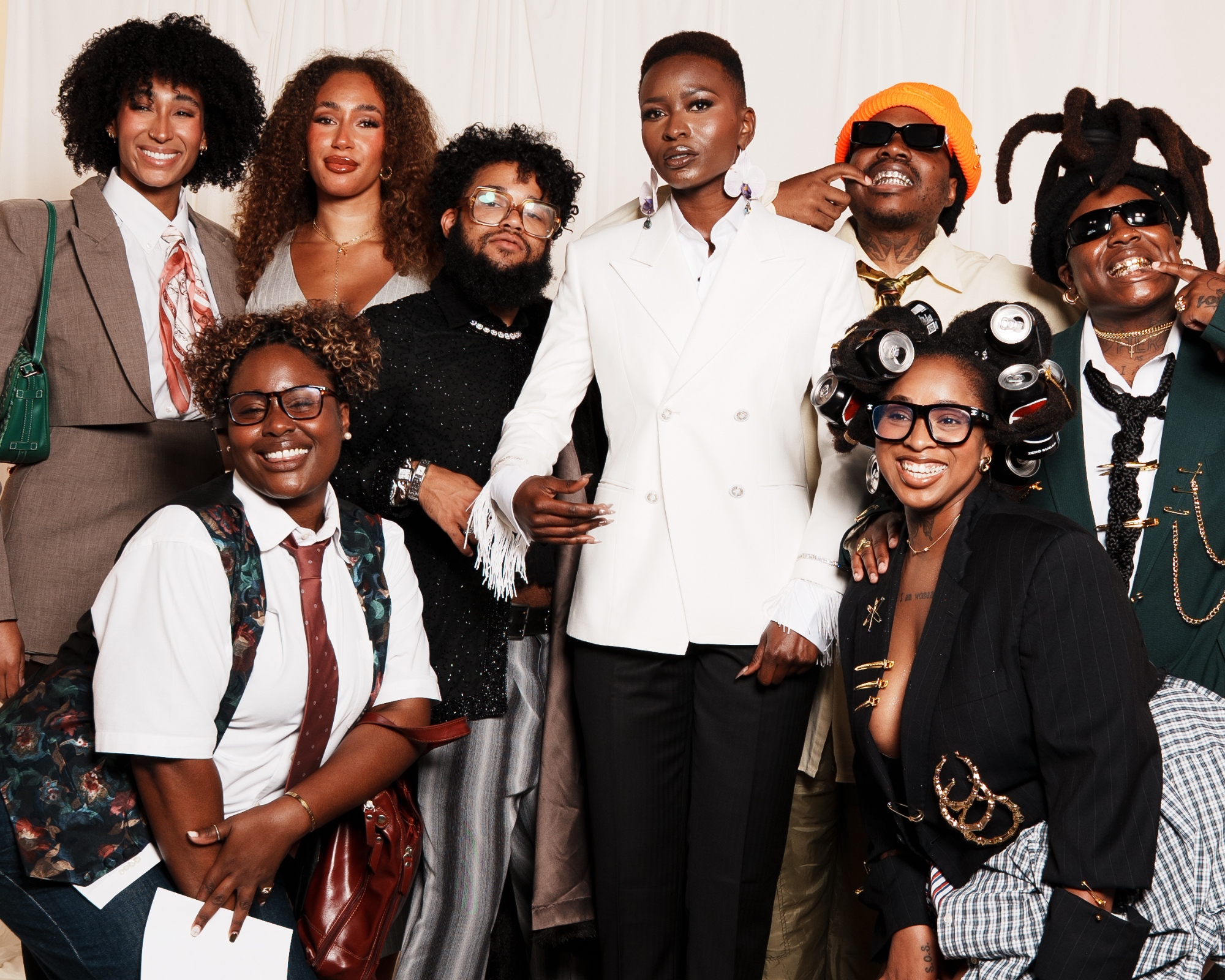 Charlotte Met Gala event. Group of eight diverse people smiling, dressed in stylish, eclectic outfits including a white suit, floral vest, and a hairdo with soda cans, posing against a plain, light background.