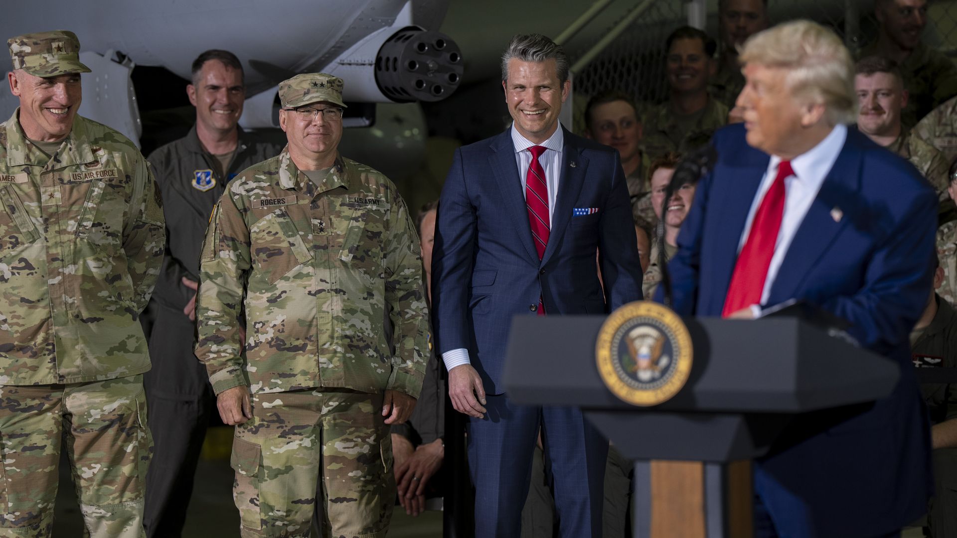 President Trump and Defense Secretary Pete Hegseth, two men in blue suits, are speaking at a military event. Soldiers surround them.