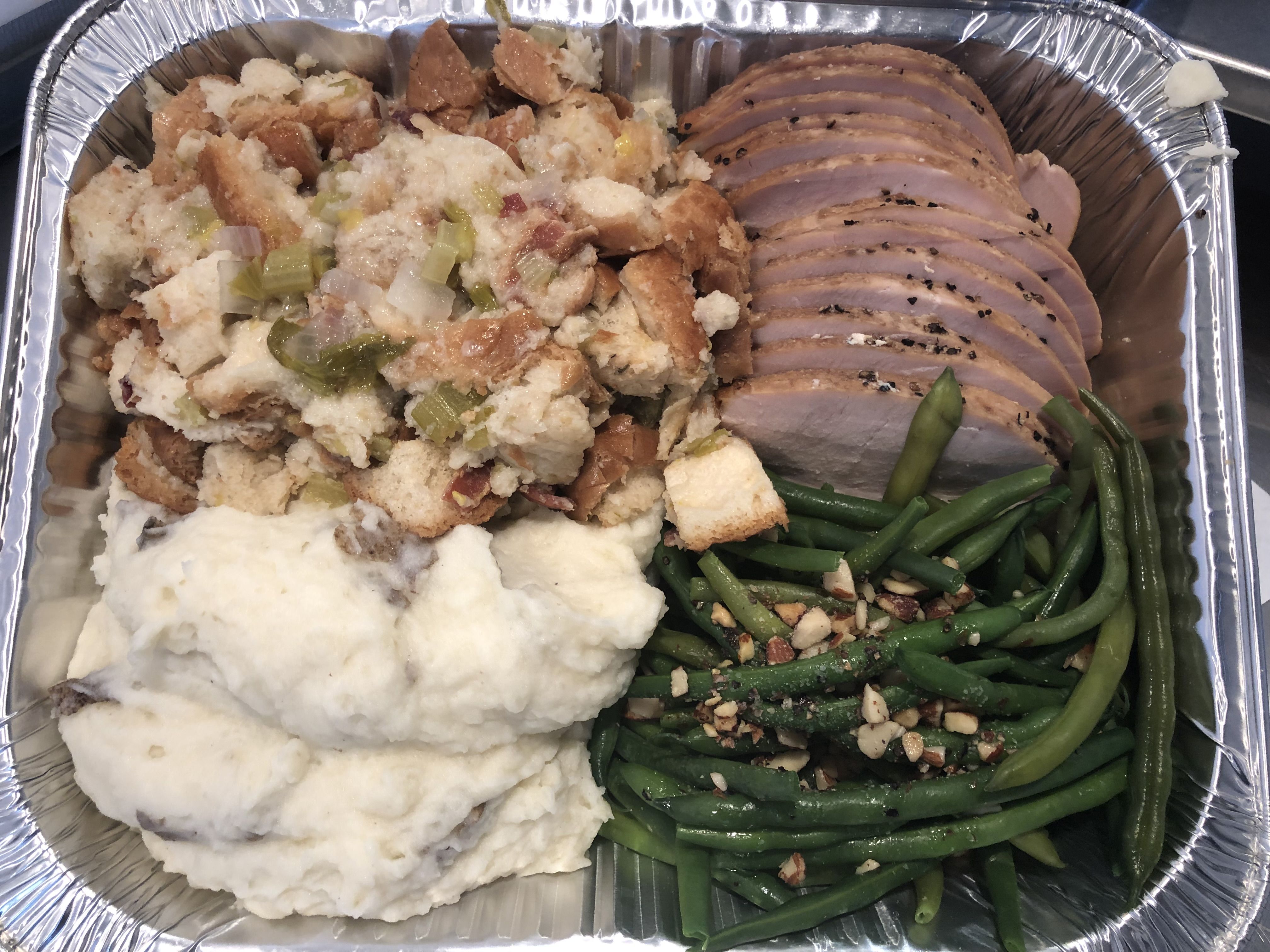 Aluminum tray with sliced roasted meat, stuffing with celery and onions, mashed potatoes, and green beans topped with chopped nuts.