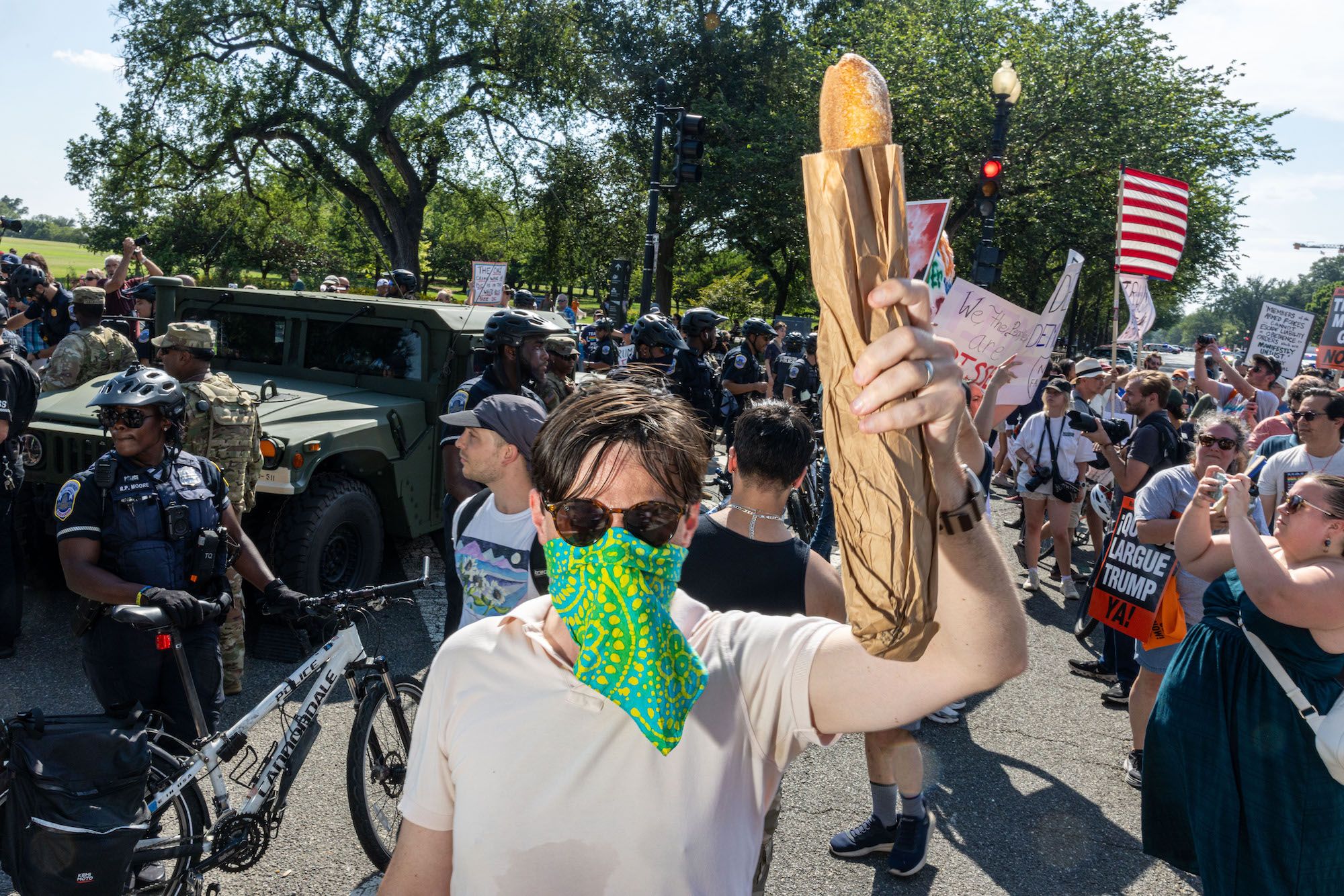 Crowd of protesters and police officers. A person with sunglasses and a green patterned mask holds a baguette wrapped in brown paper. Police in helmets and military personnel are in the background.