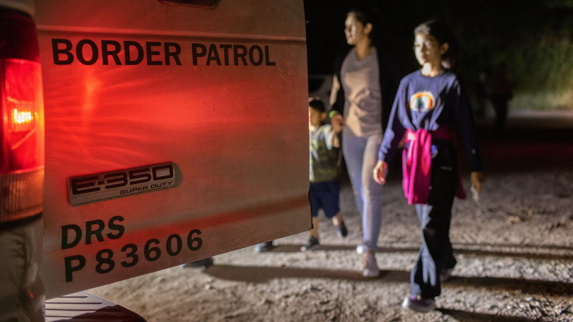 A border patrol vehicle door is illuminated as undocumented asylum seekers walk in the background