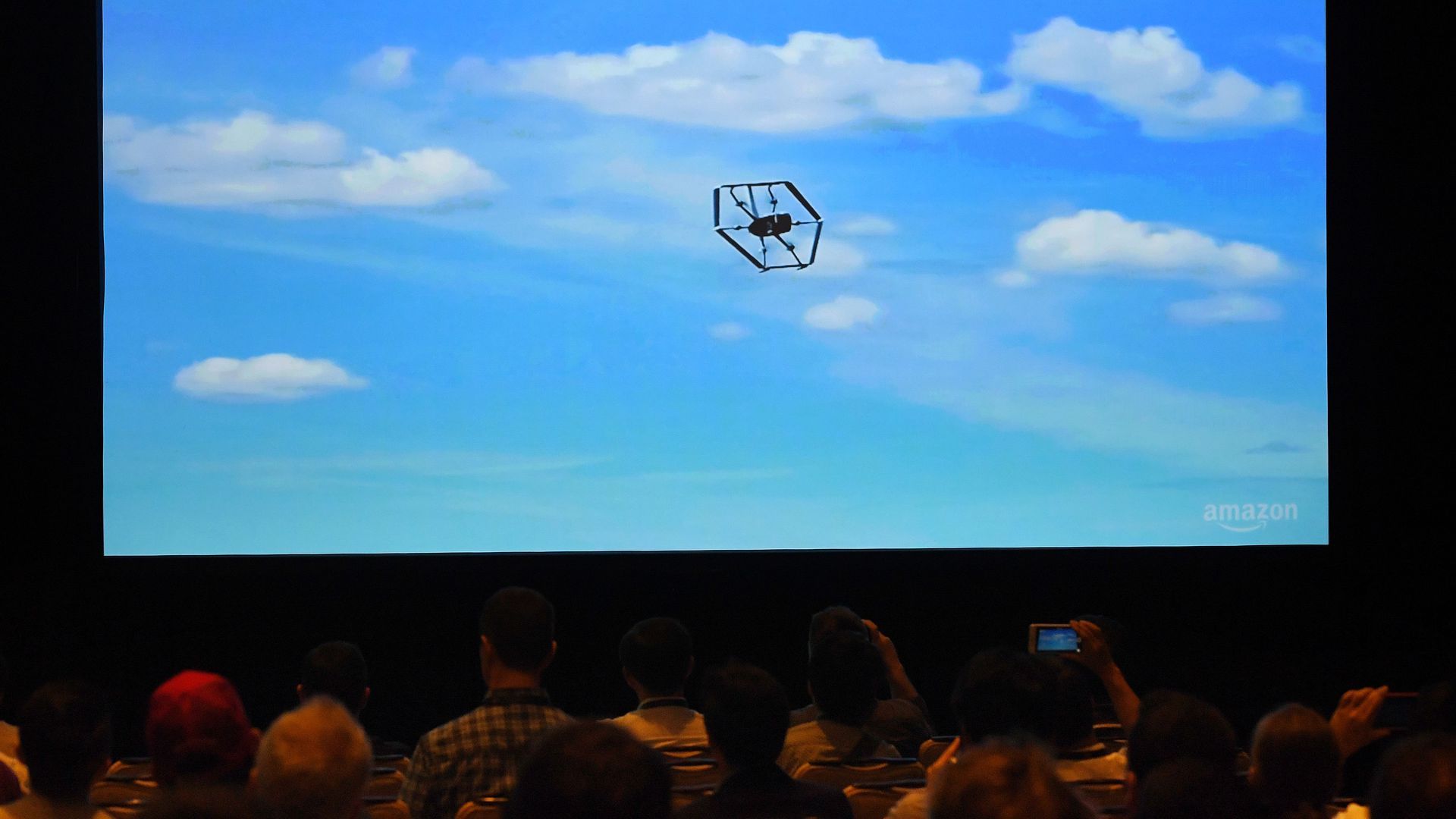 A sea of audience members look at a demo video of a drone in the sky