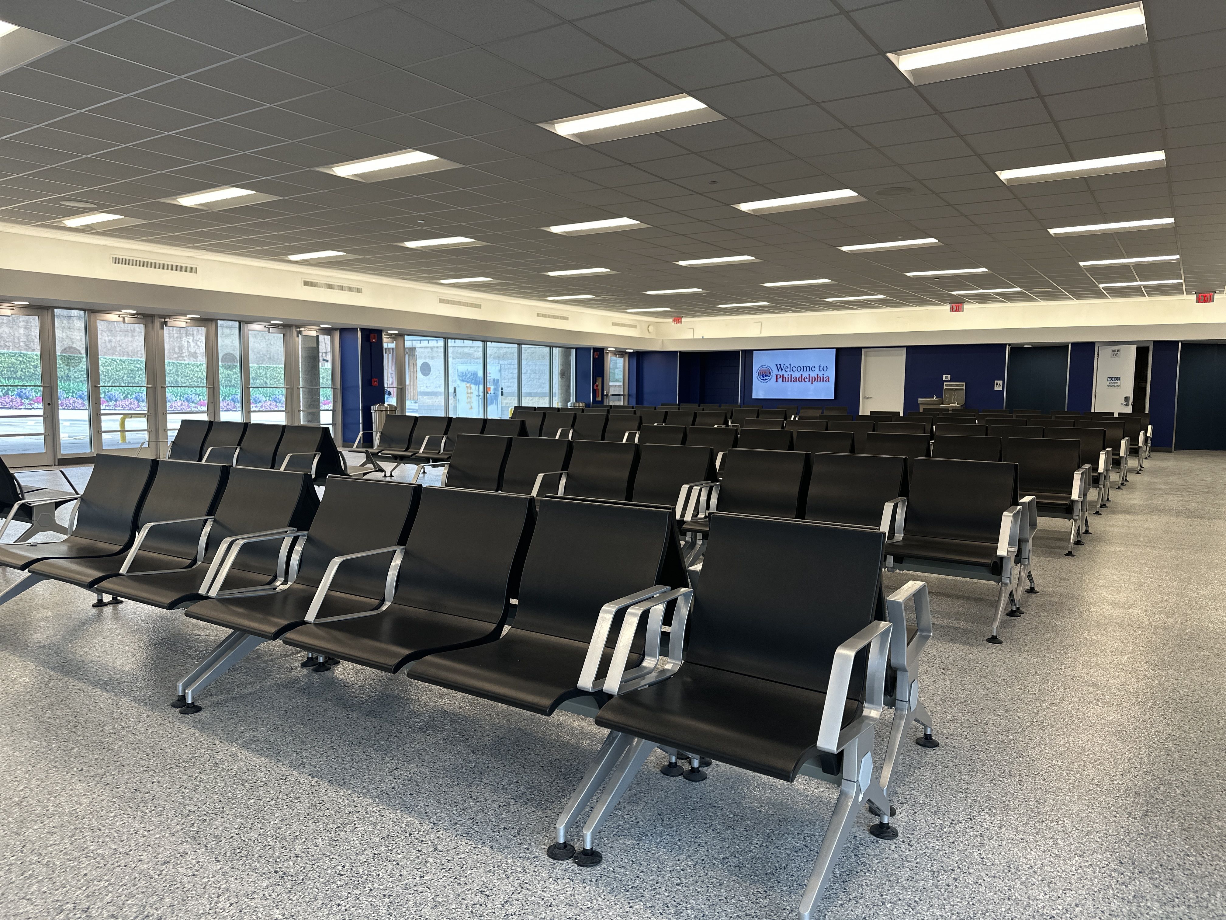 A spacious waiting area with rows of black chairs on metal frames, speckled flooring, glass entrance doors to the left, and a digital sign reading "Welcome to Philadelphia" on a blue wall.