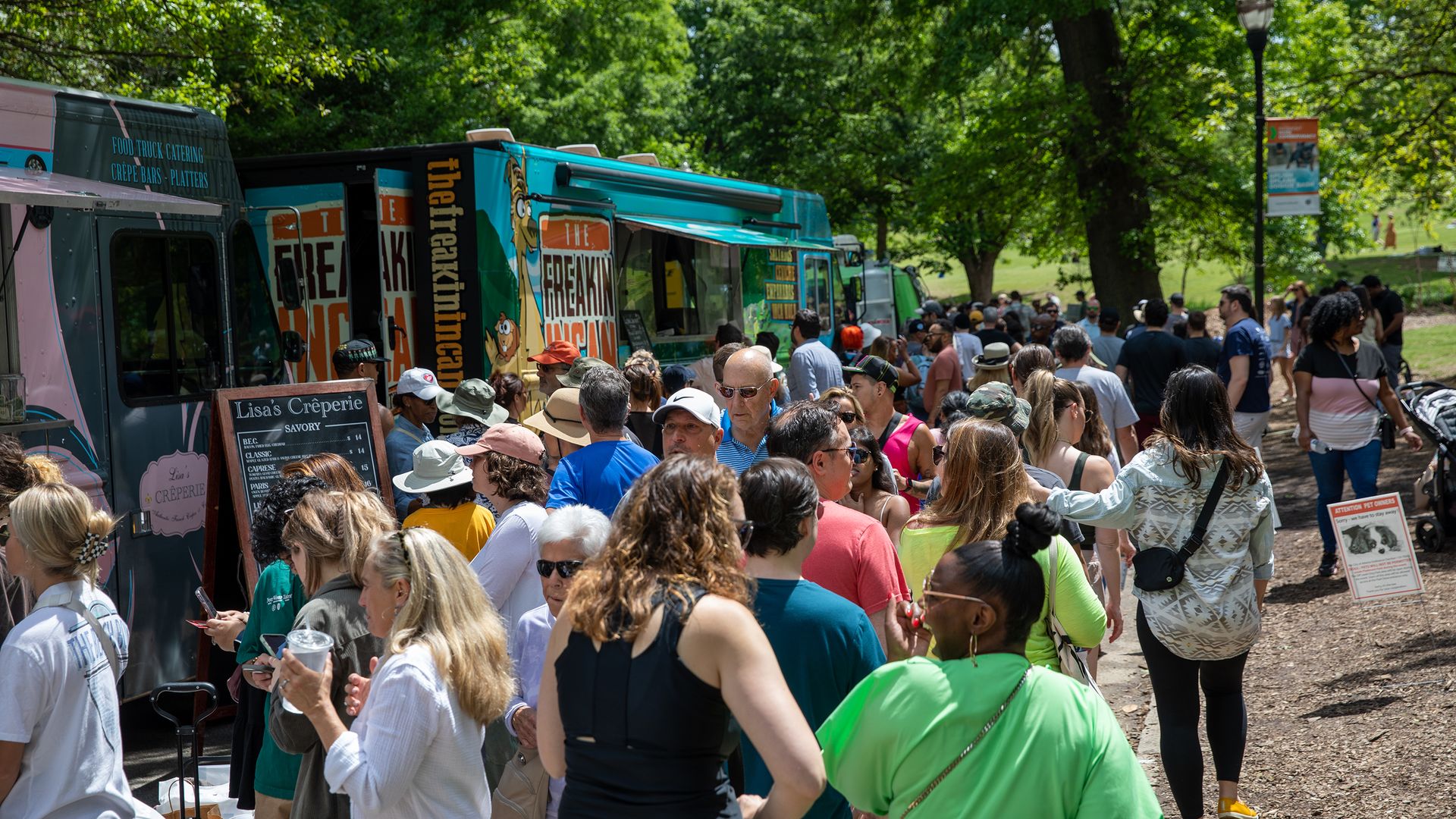Crowded outdoor food truck event on a sunny day with people in casual clothing waiting in line near trees and food trucks, including one named 