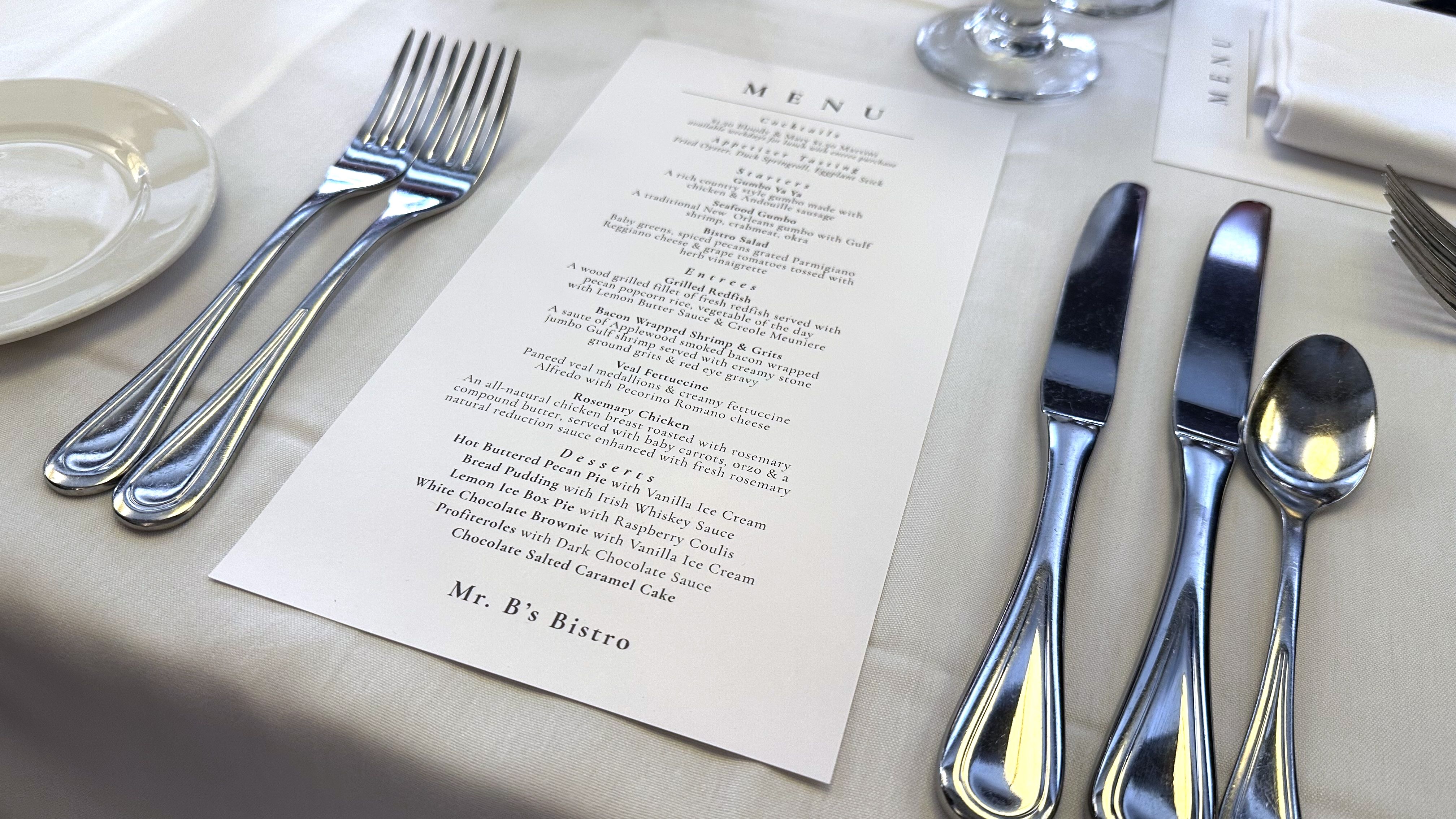 Table setting with two forks, two knives, a spoon, a small plate, and a menu for Mr. B's Bistro on a white tablecloth.