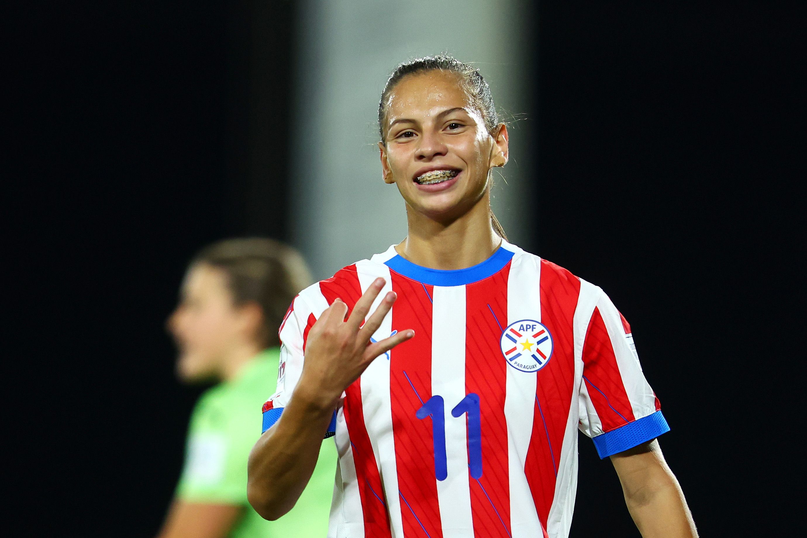 Paraguayan star Claudia Martinez in a red-and-white striped jersey with blue trim, wearing number 11 and a Paraguay crest, smiling with braces against a dark background with a blurred green-clad teammate.