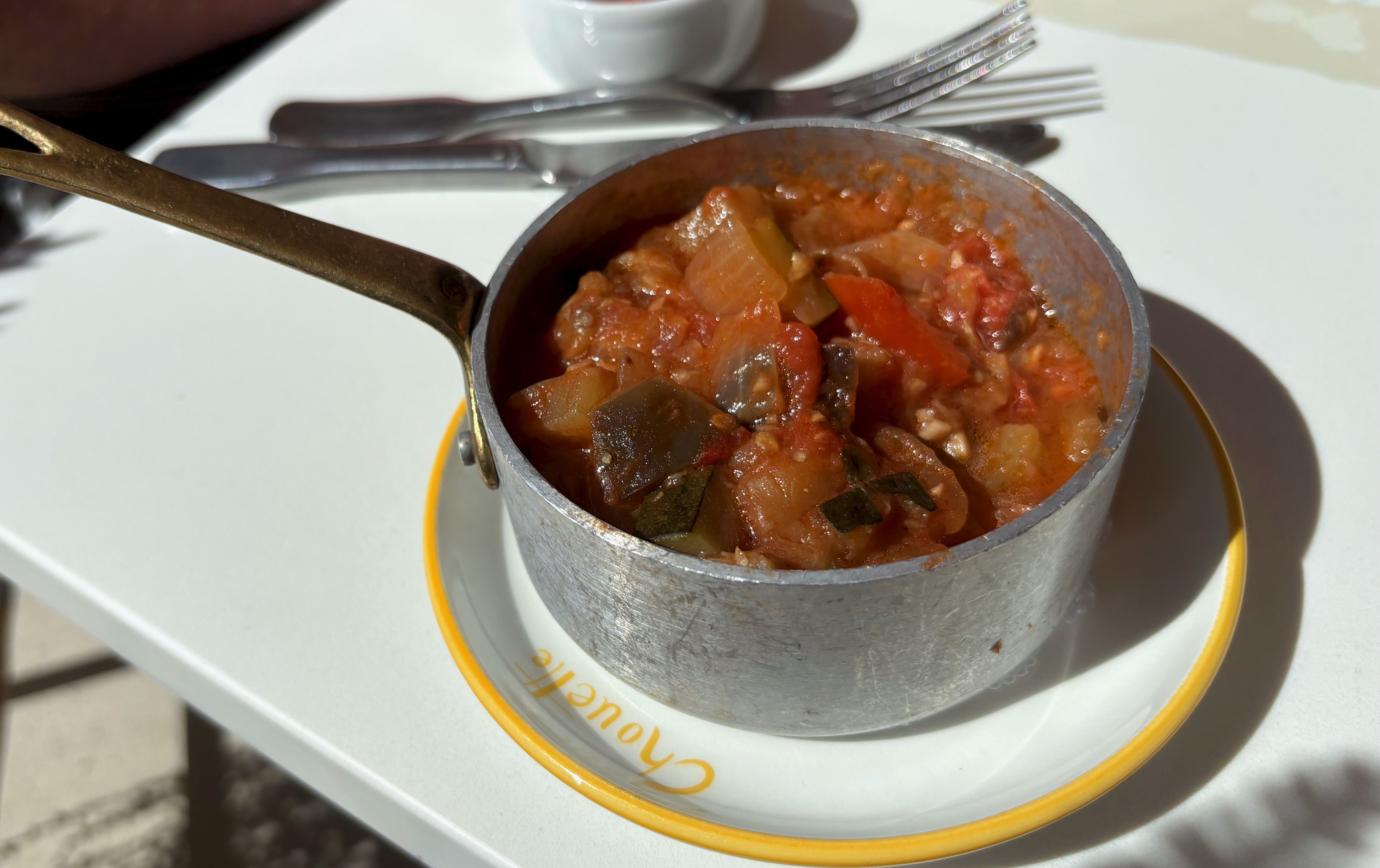 Metal saucepan with long handle filled with vegetable stew containing tomatoes, onions, zucchini, and eggplant, placed on a white plate with a yellow rim on a white table.