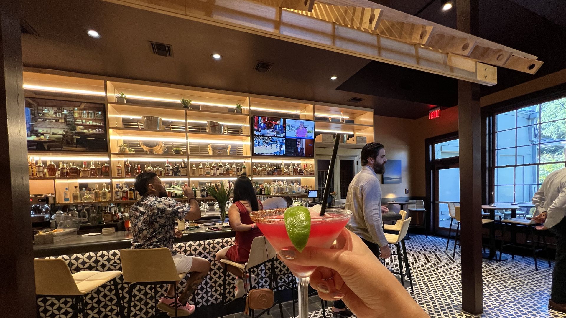 Hand holding a red cocktail with a lime wedge and black straw in a modern bar with patterned floor, patrons seated at the bar, shelves with bottles, and TV screens on the wall.