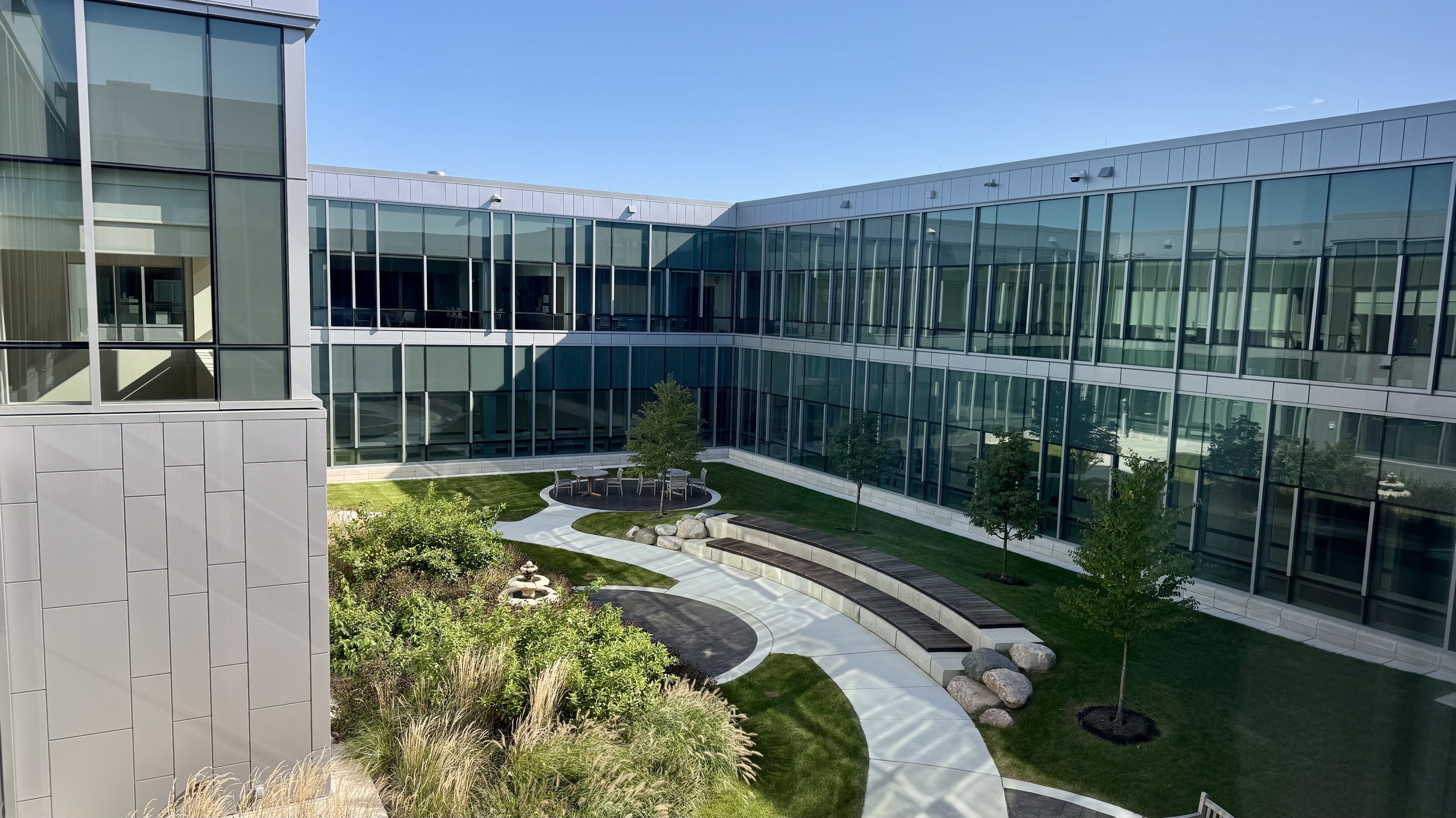 Modern glass building surrounds a courtyard with a circular seating area, round table and chairs, manicured lawn, trees, and stone landscaping; blue sky overhead.
