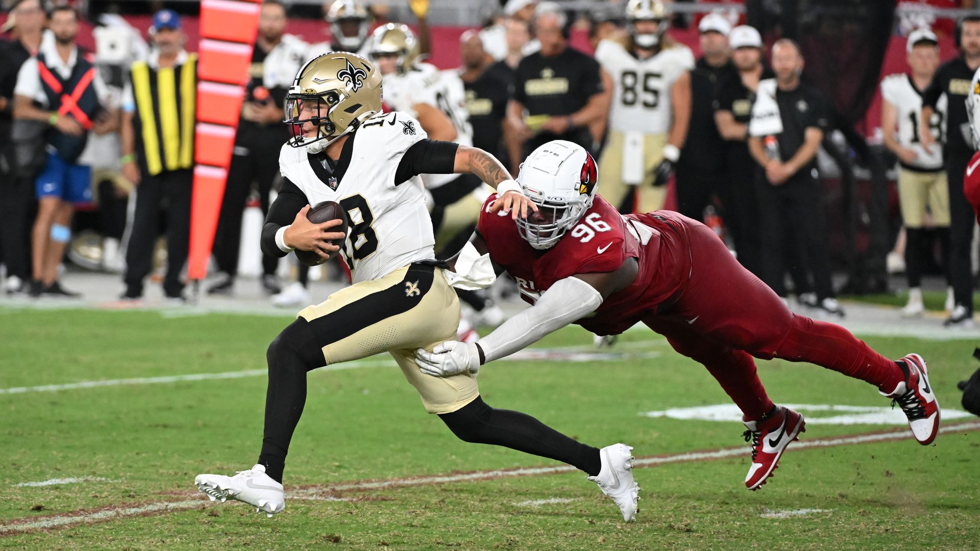 A football player in a white and gold New Orleans Saints uniform runs with the ball while a player in a red Arizona Cardinals uniform lunges to tackle him on a green field during a game.