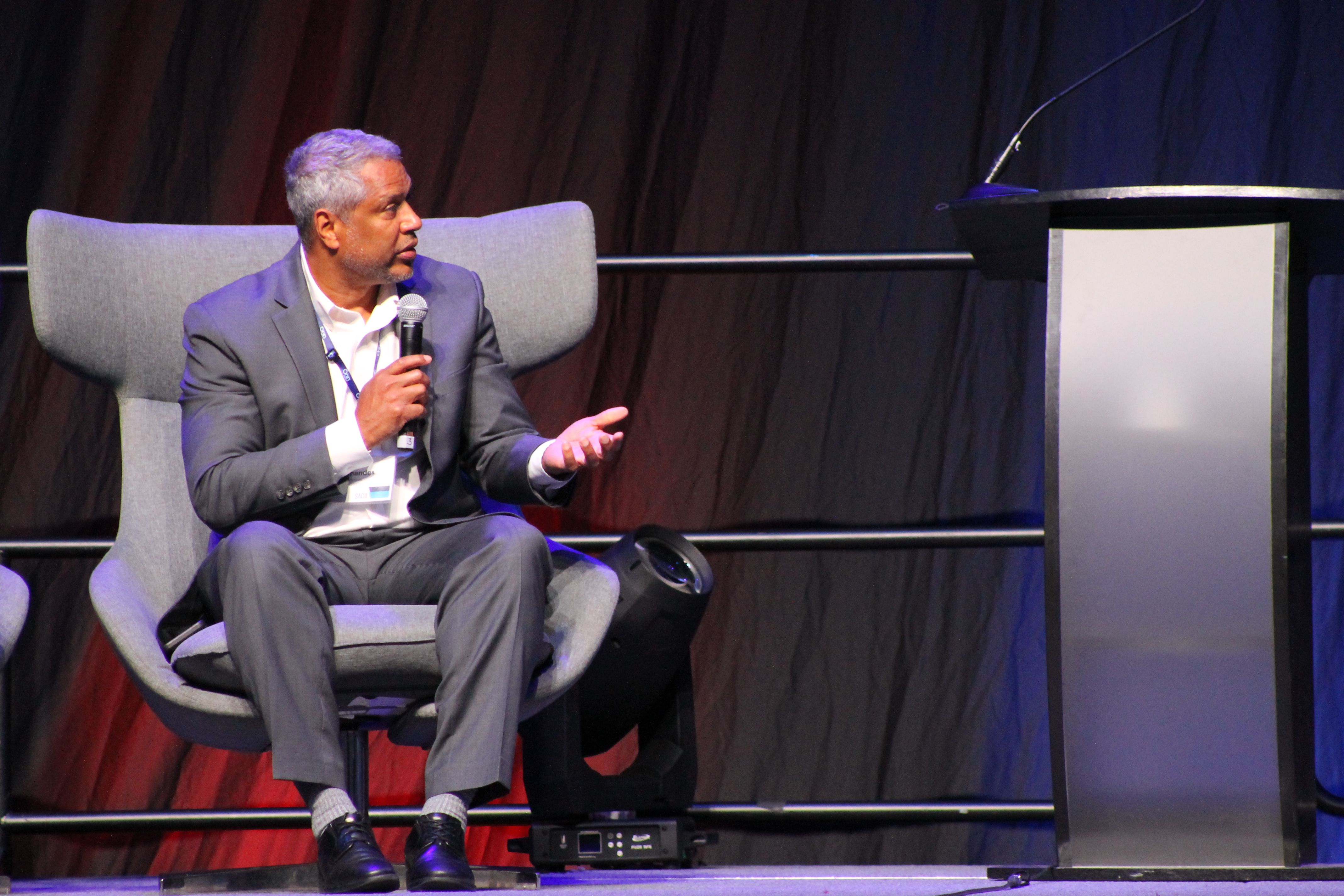 Man in gray suit holding microphone and gesturing while seated on stage in gray chair next to a podium and black backdrop with blue and red lighting.