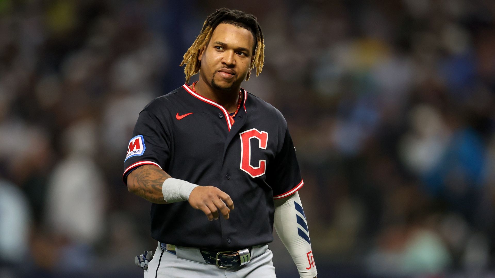 Baseball player in a dark gray uniform with red trim and a large red C on the chest, with blond dreadlocks and a wrist wrap, on a field with a blurred crowd behind.