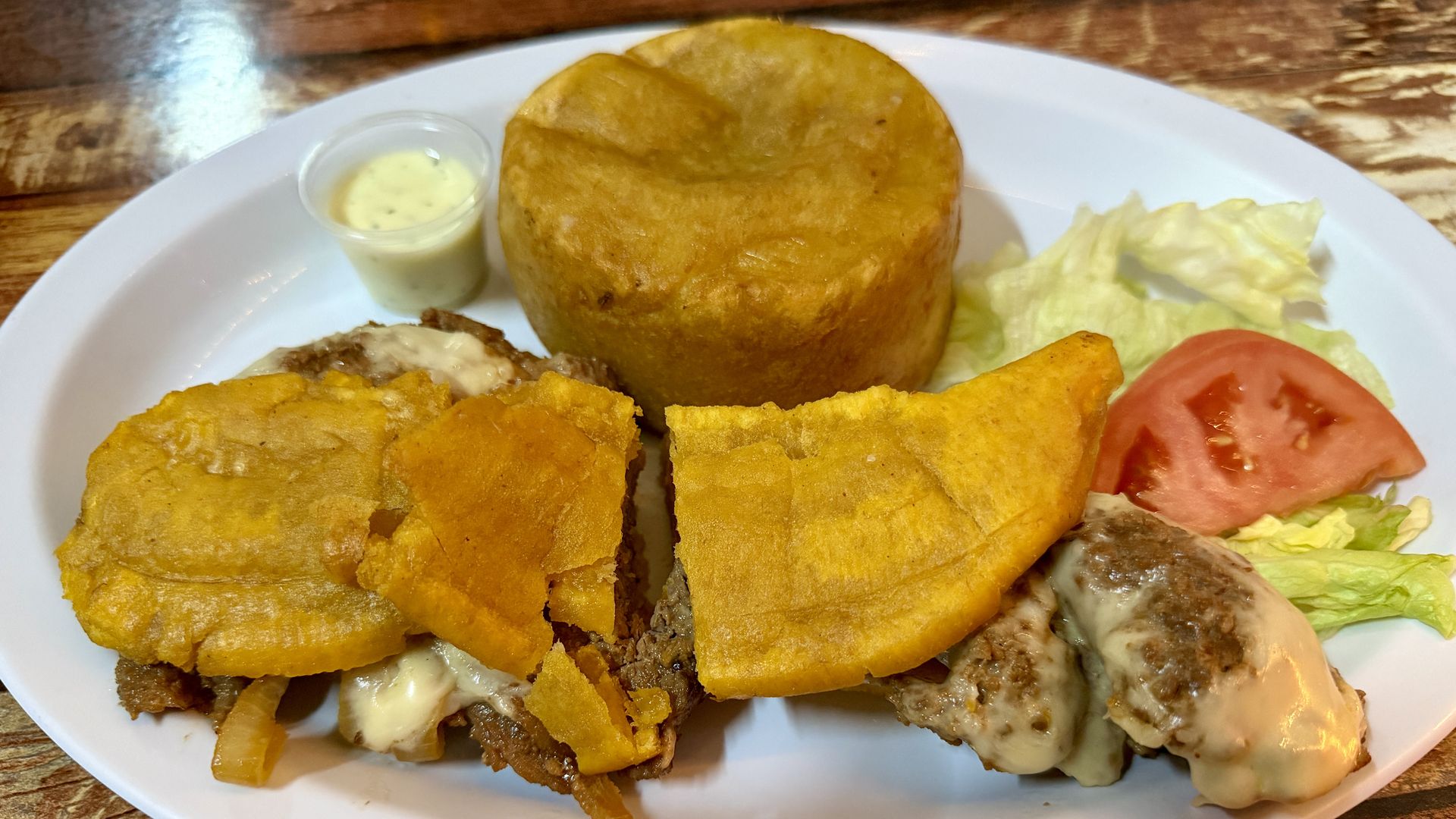 Plate with fried meat topped with cheese and fried plantain slices, a mound of fried cornmeal, lettuce, tomato slice, and a small container of dipping sauce on a wooden table.