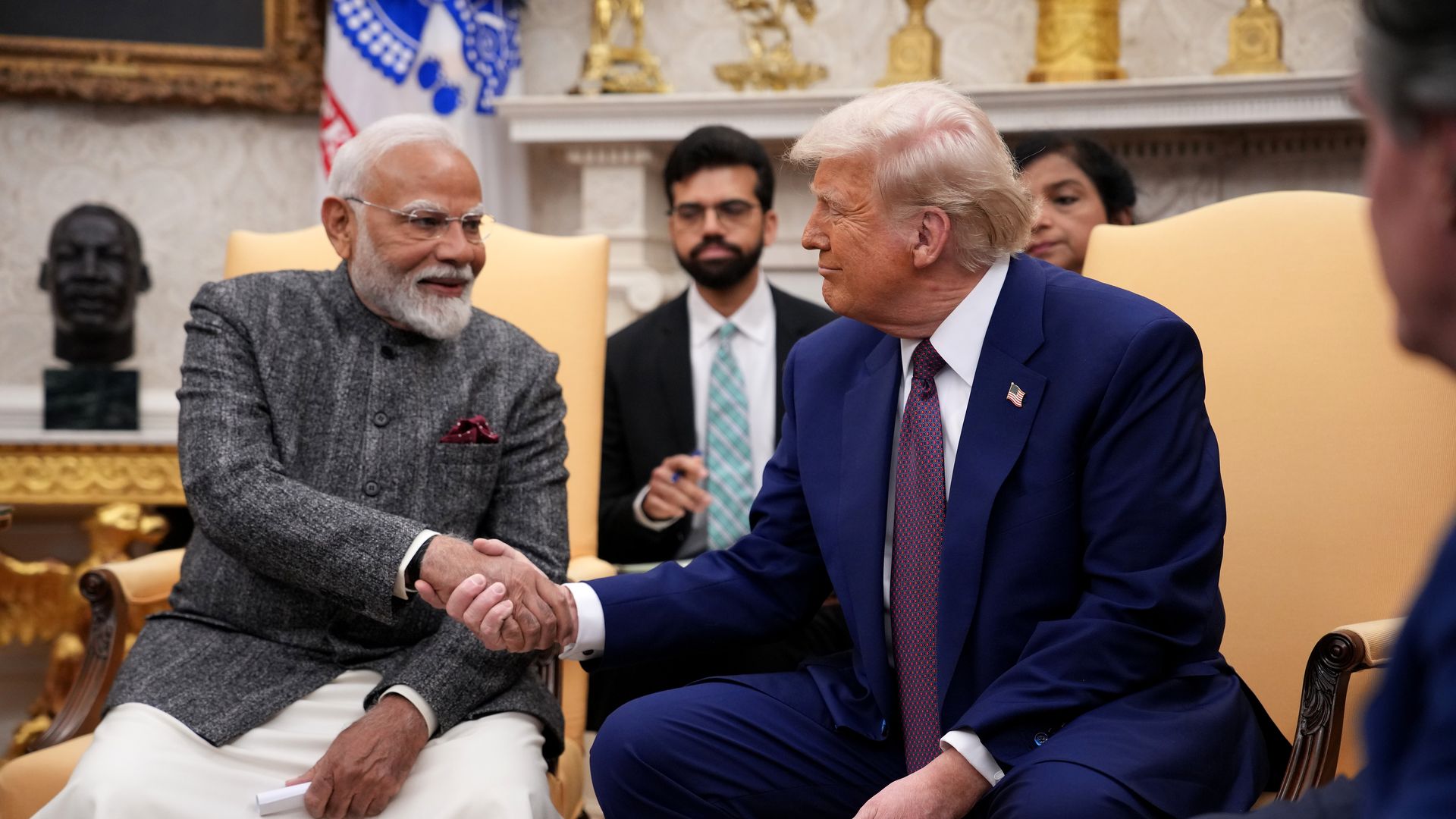 India's Prime Minister Modi in gray jacket and white pants shakes hands with U.S. President Trump in blue suit and red tie, seated in a decorated room with yellow chairs and officials in background.