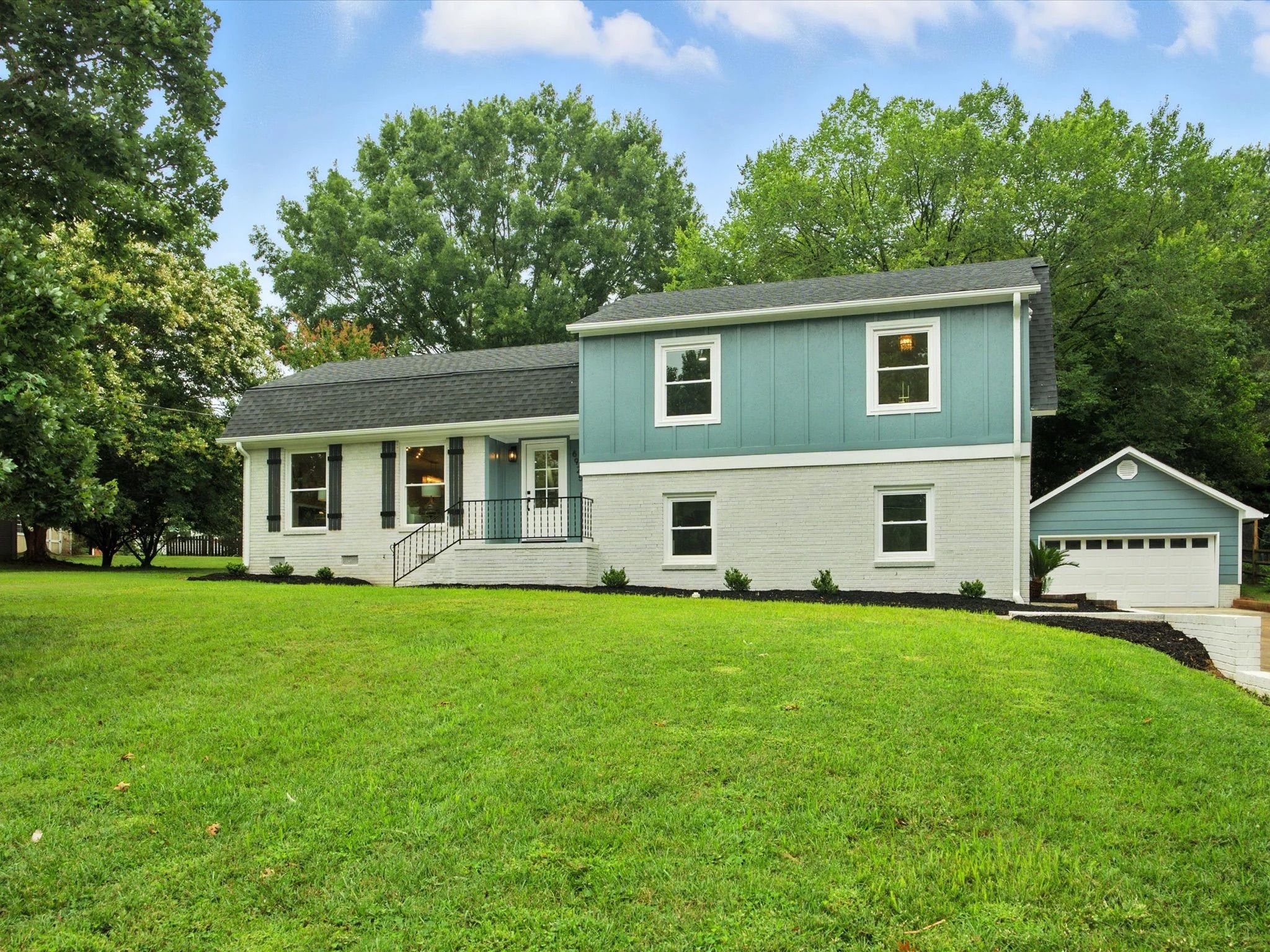 Two-story house with white brick on lower level and blue paneling on upper, black roof, black window shutters, a green lawn, trees, and a detached garage to the right under a blue sky.