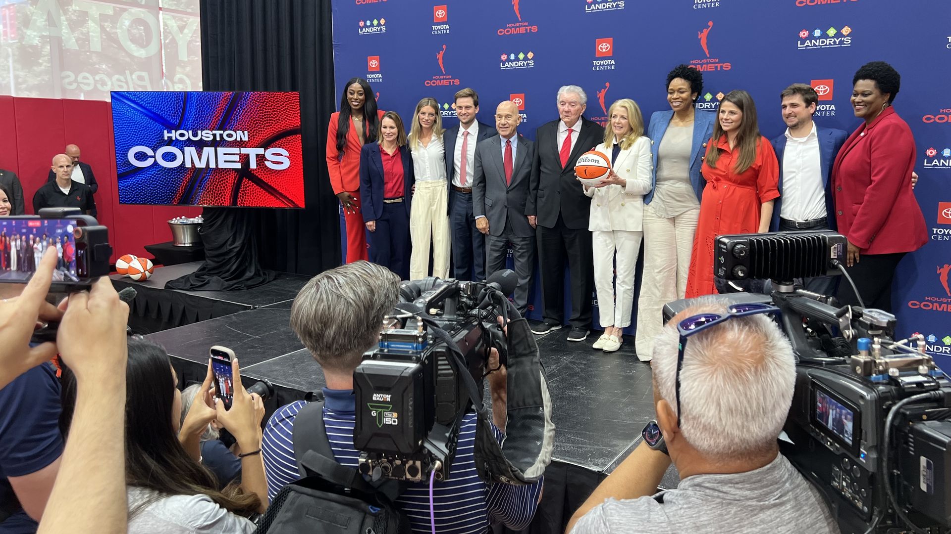 Group of people posing on a stage in front of a blue backdrop with Houston Comets logos; a woman in a white suit holds a basketball, others in red and business attire; photographers in the foreground.