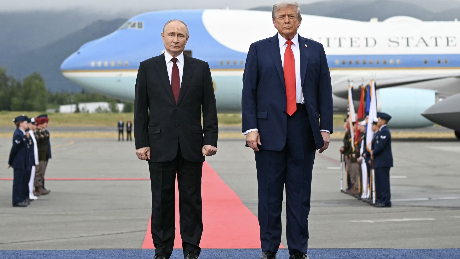 Two men in suits stand on a blue carpet with a red carpet behind them at an airport; a large Air Force One plane is in the background, with military personnel lined up on each side.