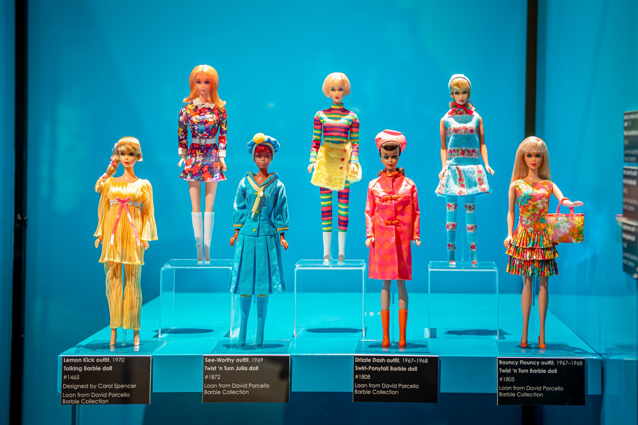 Eight vintage Barbie dolls in bright, patterned outfits stand on clear pedestals in a glass display against a vivid blue backdrop; plaques label each outfit.
