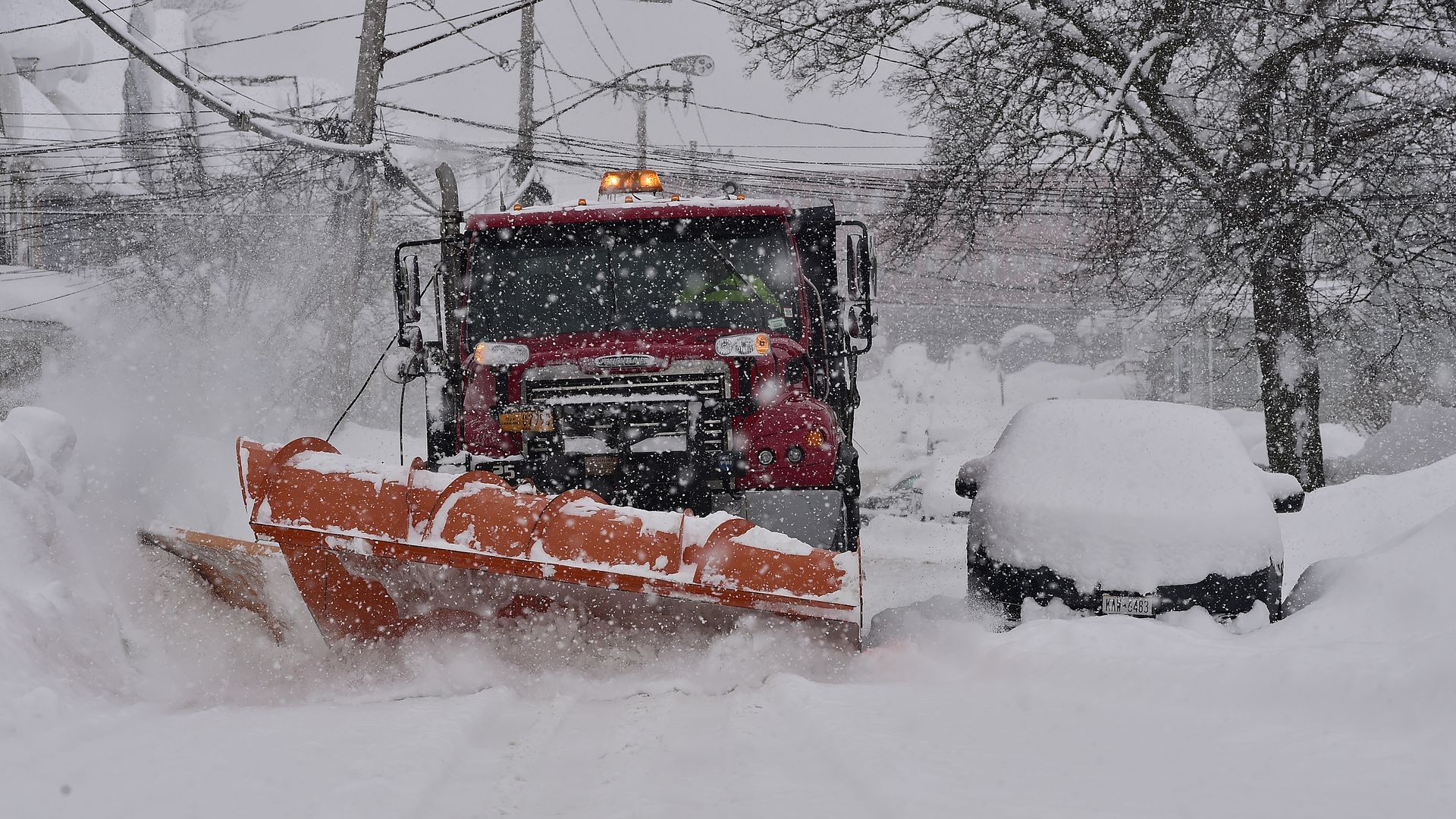 A red snowplow truck driving through piles of snow near a car covered with snow.