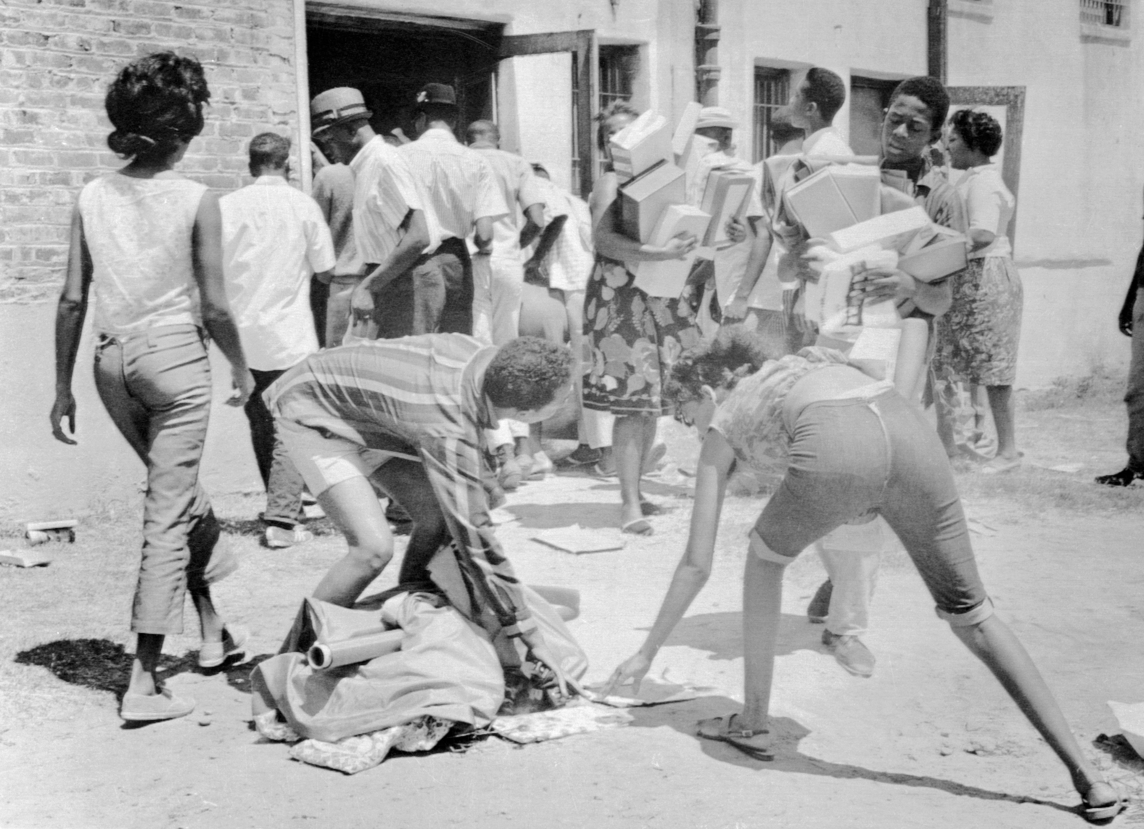 Black-and-white photo of people outside a building; two individuals bending to pick items from the ground, others carrying boxes and standing in line near an entrance.