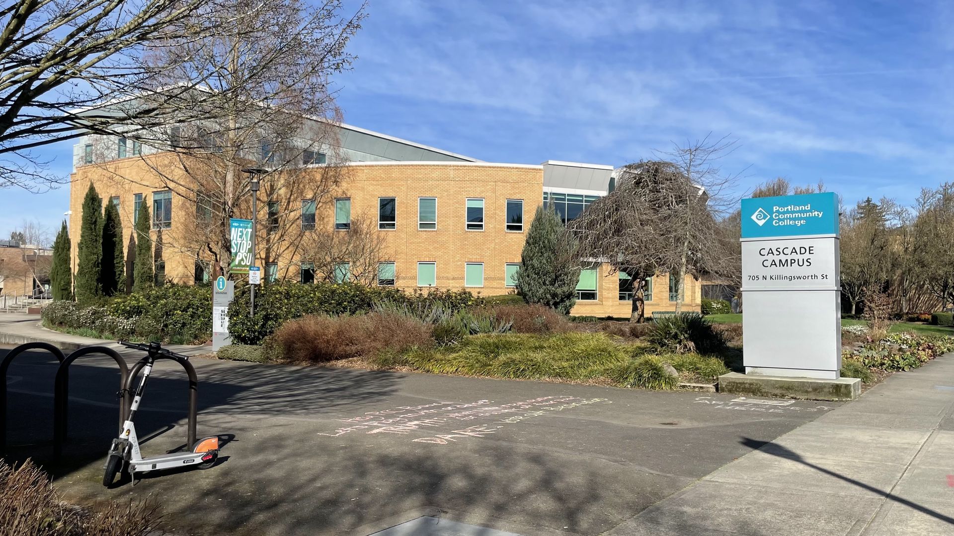 Portland Community College Cascade Campus sign beside a tan brick building under a blue sky; a gray scooter rests by bike racks in the foreground, with trees and shrubs around.