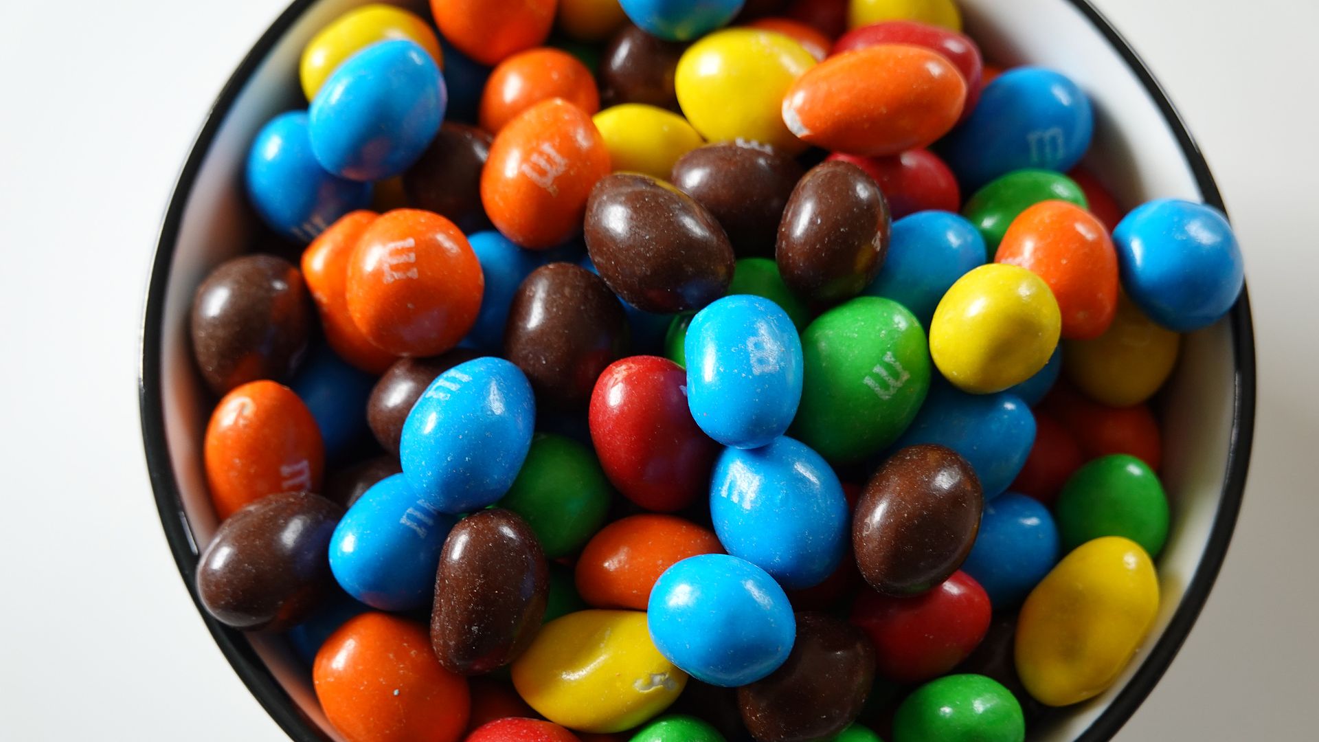 Close-up of a white bowl filled with colorful candy-coated chocolates (M&M-like) in blue, red, orange, yellow, green, and brown.