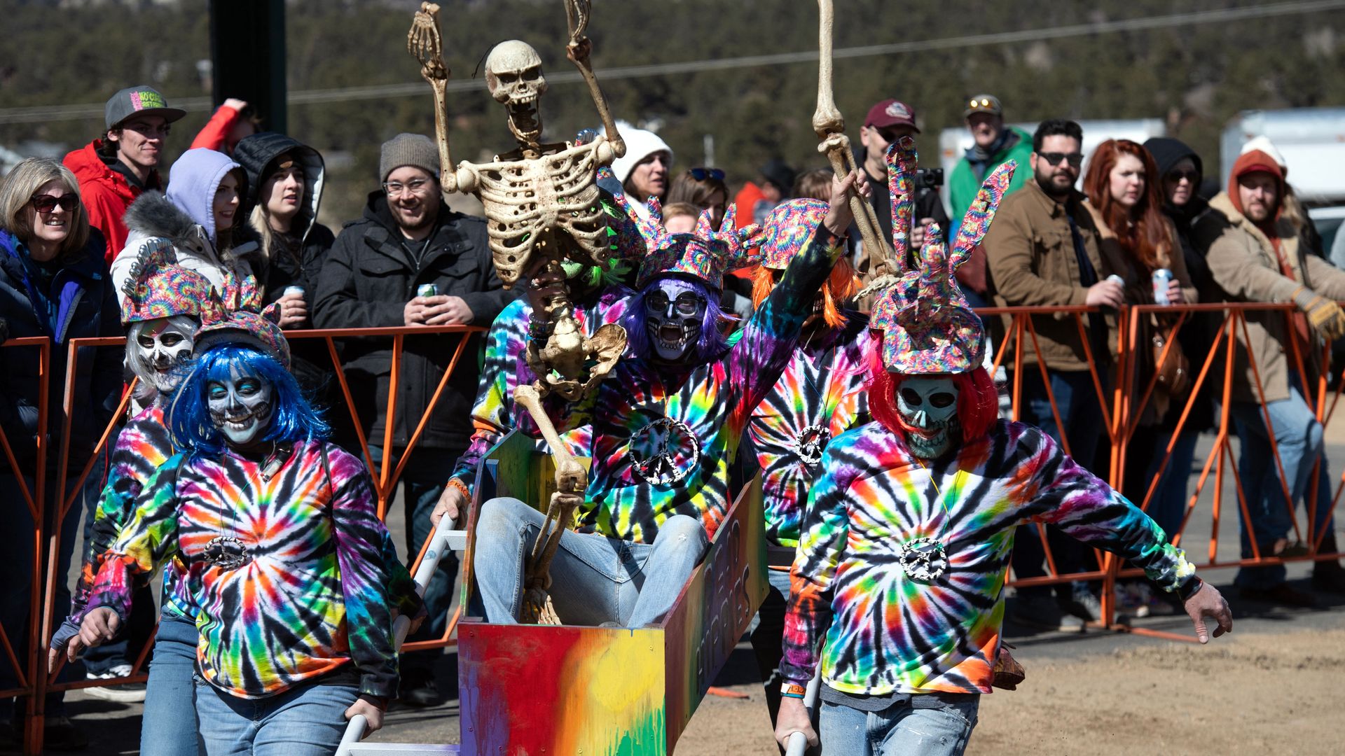 Colorful parade scene with performers in tie-dye shirts and skeleton masks, carrying a rainbow-painted float while spectators stand behind orange barriers.