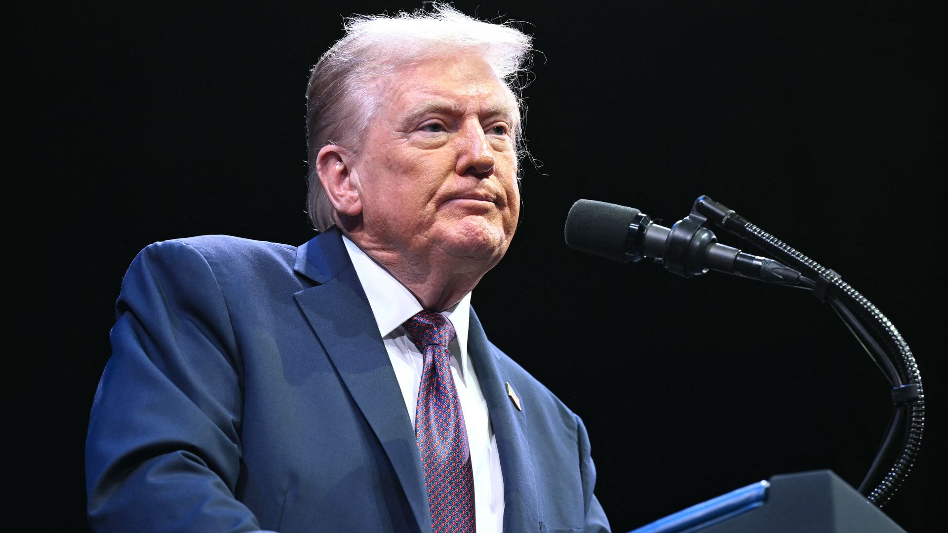 President Trump, wearing a navy jacket with a small US flag pin on it, a maroon and blue check tie and a white shirt, frowns while pausing from speaking at a mic.