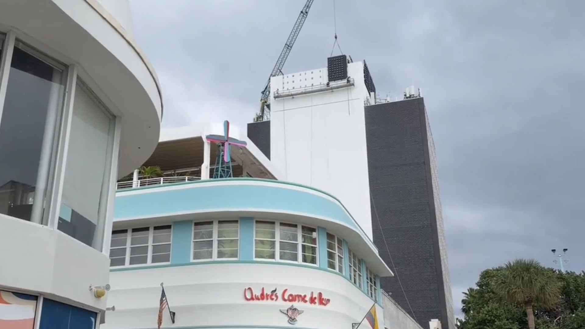 Curved white and light blue building with windows, signage "Andrés Carne de Res", flags, and palm trees, beneath a cloudy sky and a tall dark and white tower with a crane above.