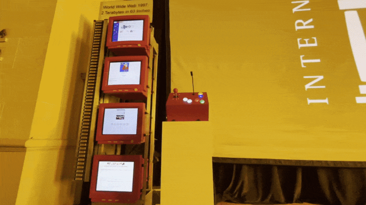 Stack of four red-framed monitors displaying early World Wide Web pages, labeled 'World Wide Web 1997 2 Terabytes in 69 inches' on yellow wall next to a podium with controls.