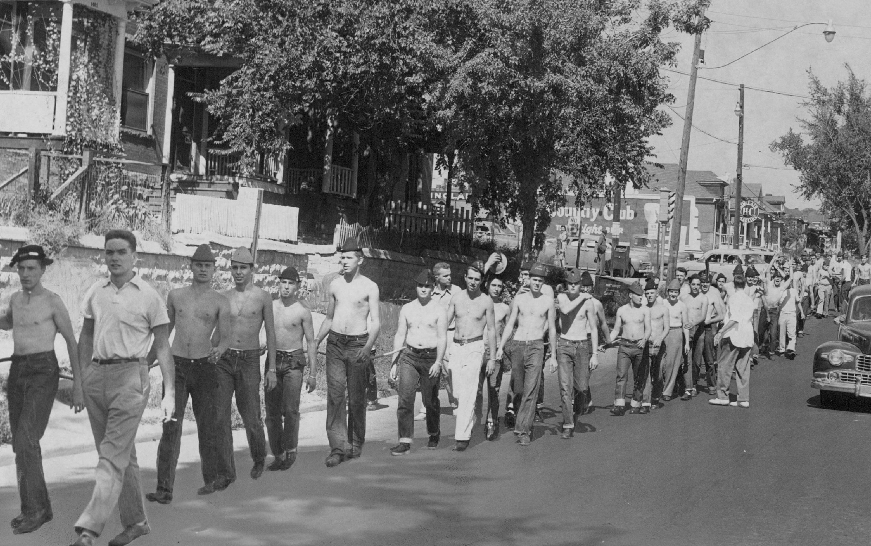 A black-and-white photo of men, some shirtless, walking down the street in a single file.