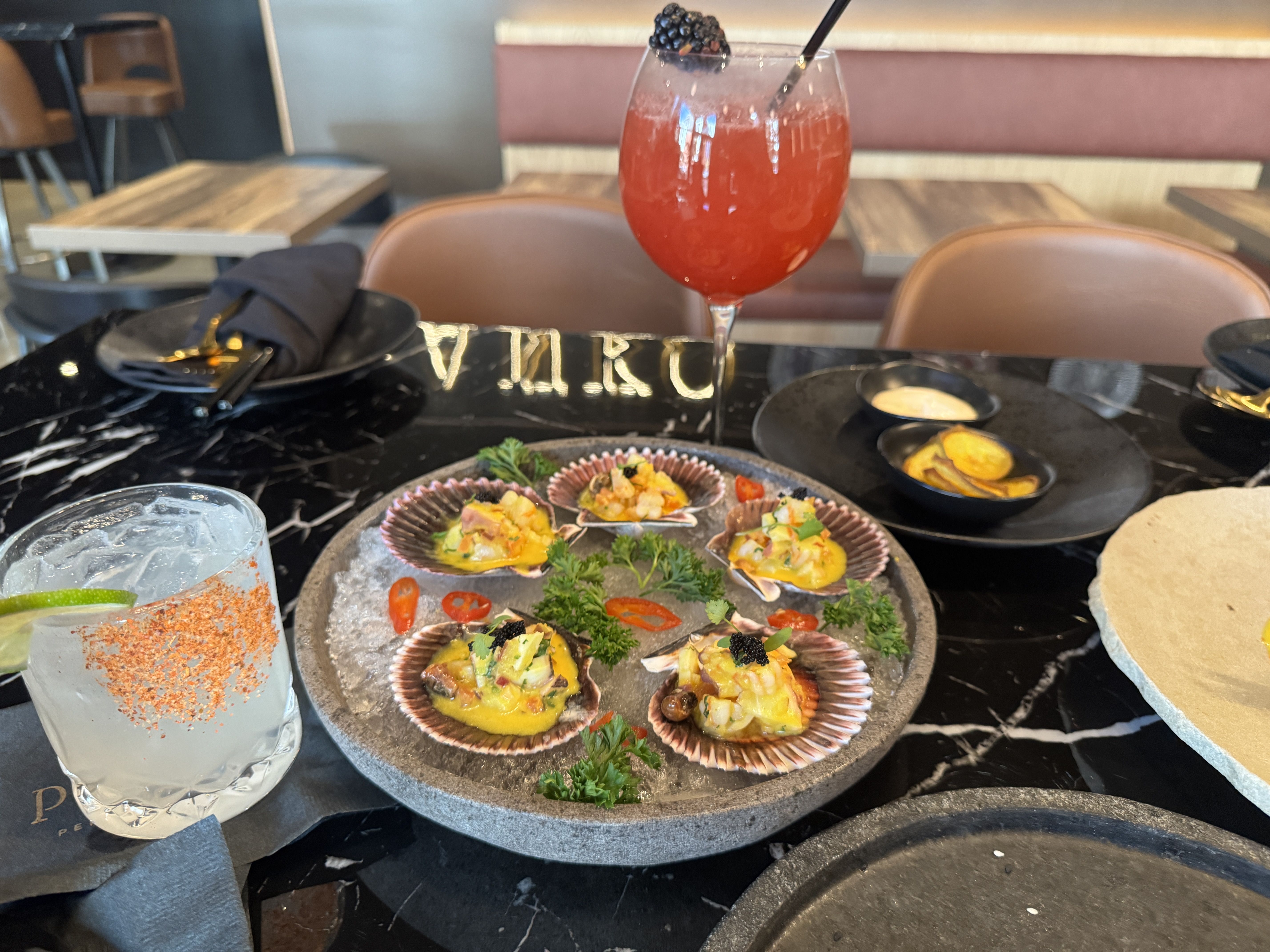 Table with a plate of five scallop shells filled with seafood ceviche, garnished with herbs and sliced red chili, an icy margarita glass with chili salt rim, and a tall red cocktail with a blackberry.