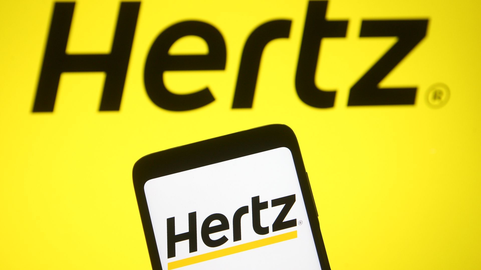 Picture of a phone screen with the Hertz logo on it. The phone is placed in front of a wall with the Hertz logo also on it.